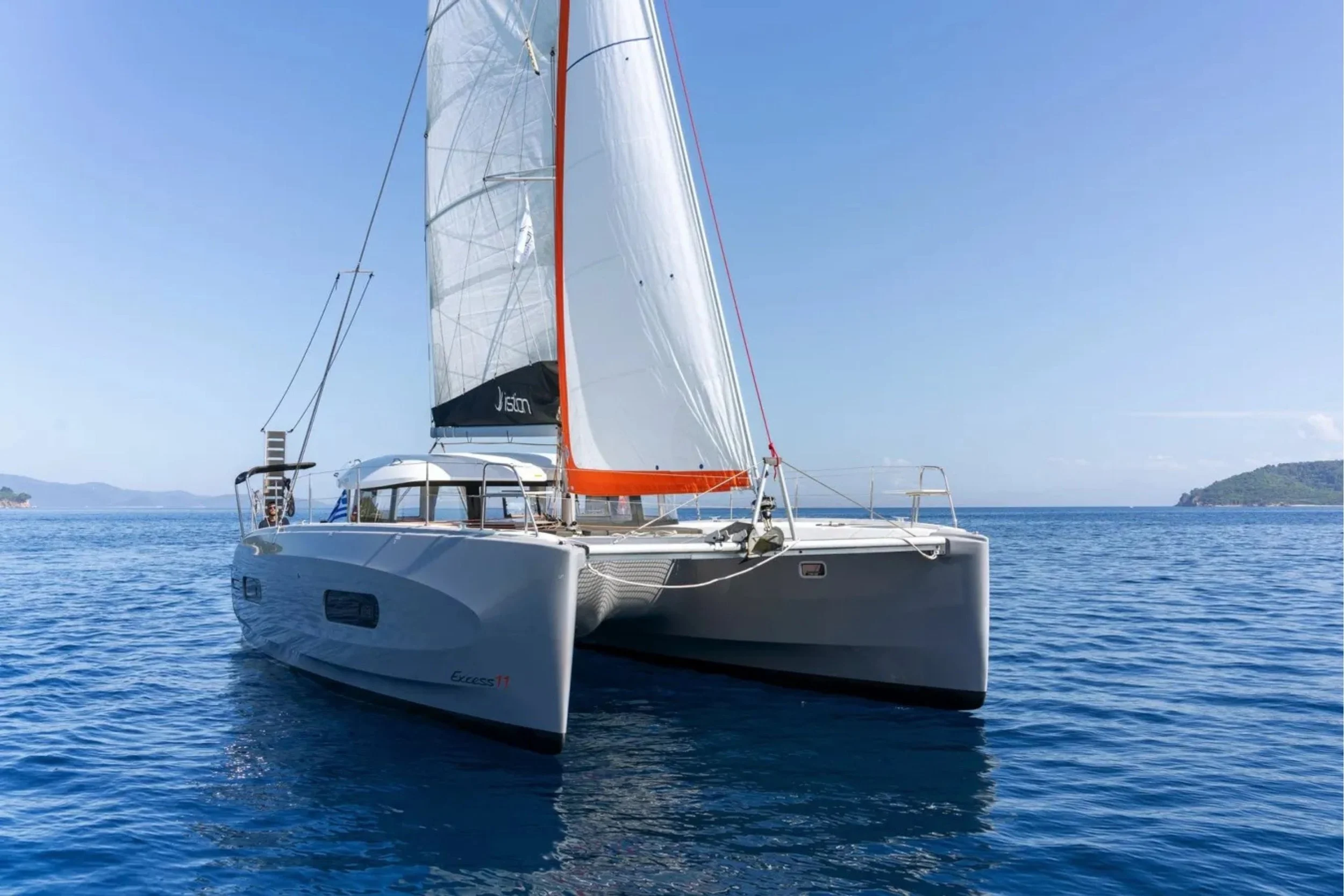 A white sailing catamaran with large sails on calm blue ocean water, distant land on the horizon under clear skies.