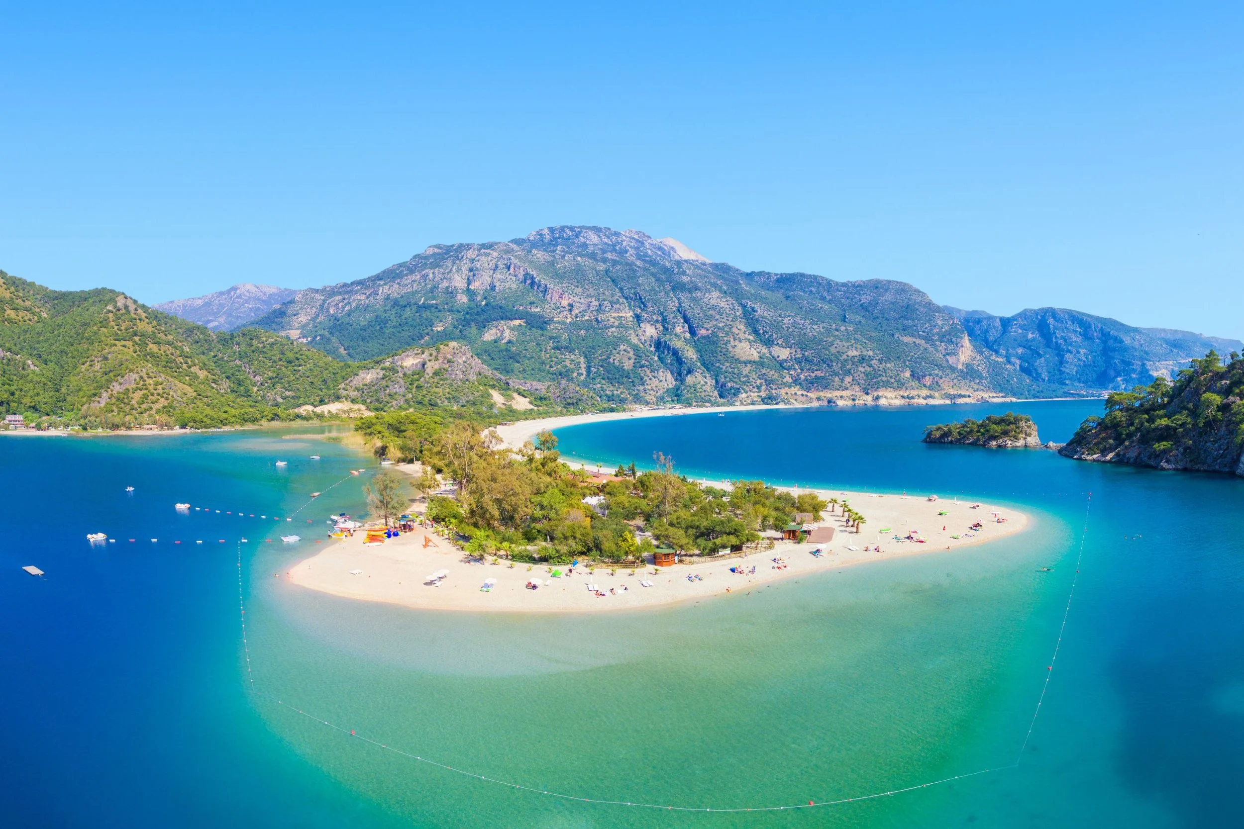 An aerial view of a beach with a sandbar surrounded by calm turquoise water, lush green trees, and mountains in the background under a clear blue sky.