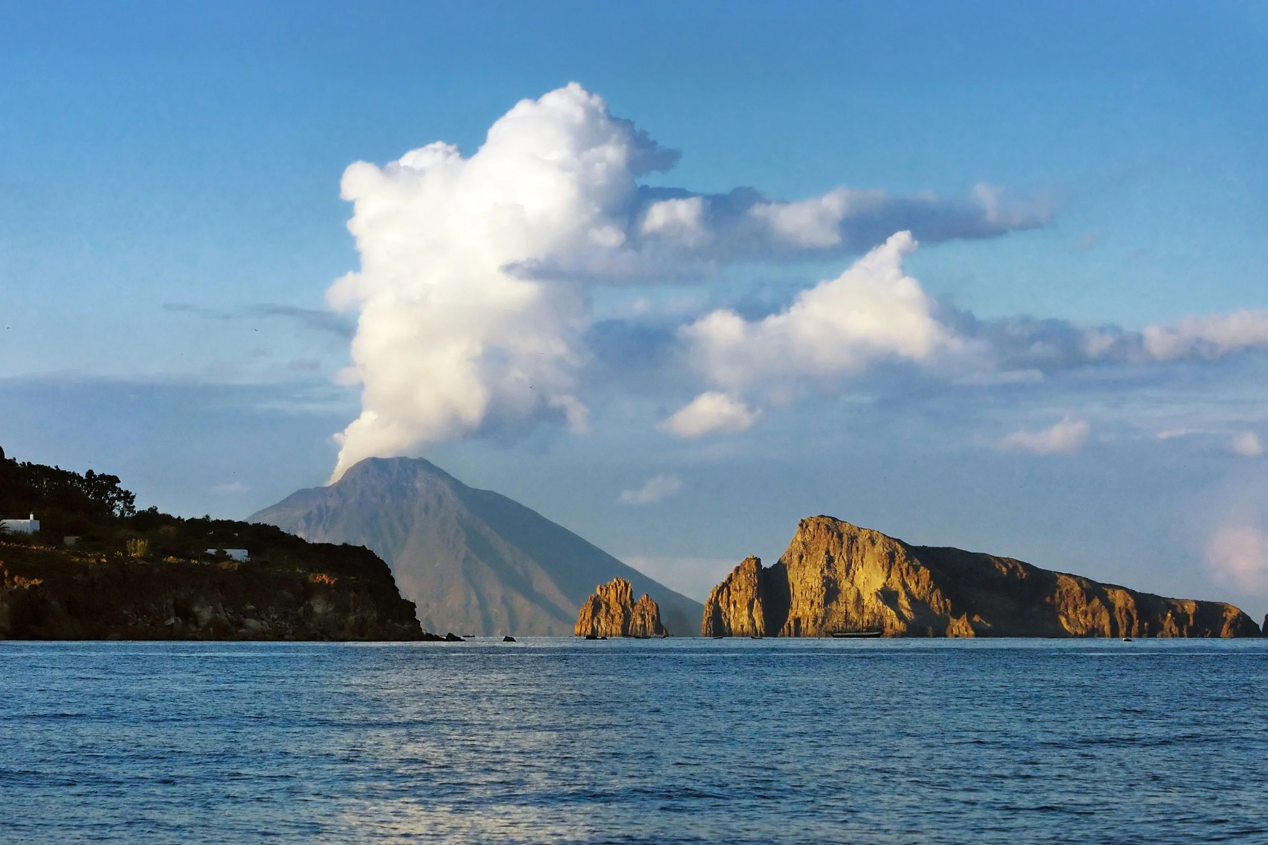 Ocean with rocky cliffs and tree-covered land, erupting volcano in background emitting smoke, blue sky with fluffy clouds.