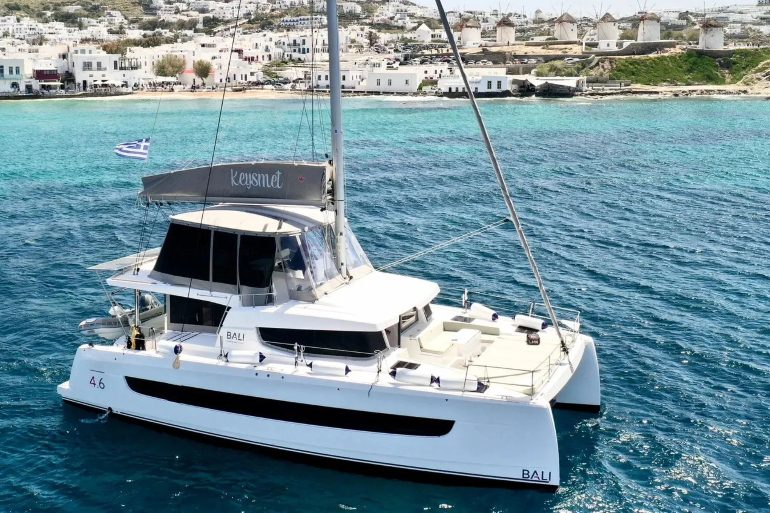 A white yacht named 'BALI' sailing in turquoise waters near a coastal town with white buildings and windmills in the background. A Greek flag is flying on the yacht.