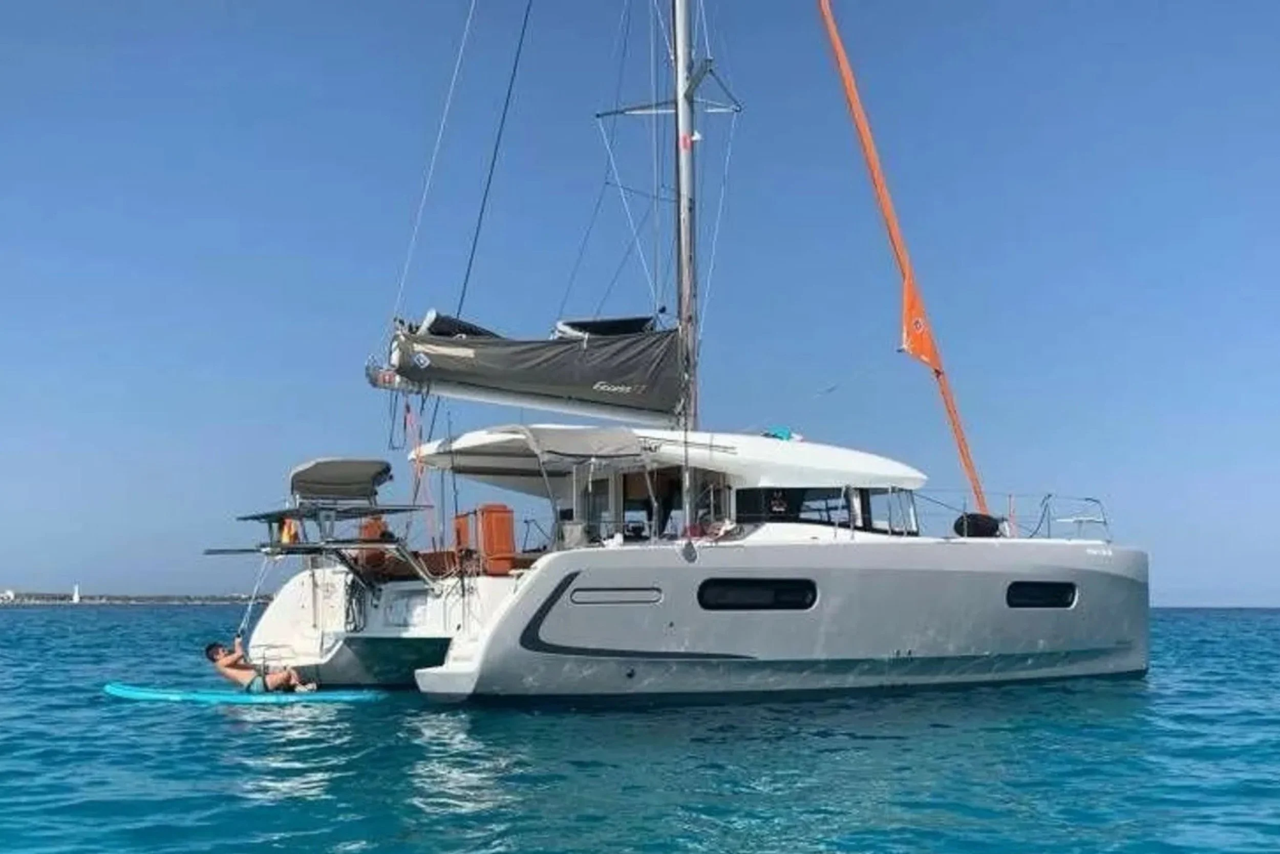 A white sailboat with black sails floating on clear blue water under a bright blue sky, with a person lying on a paddleboard beside it.