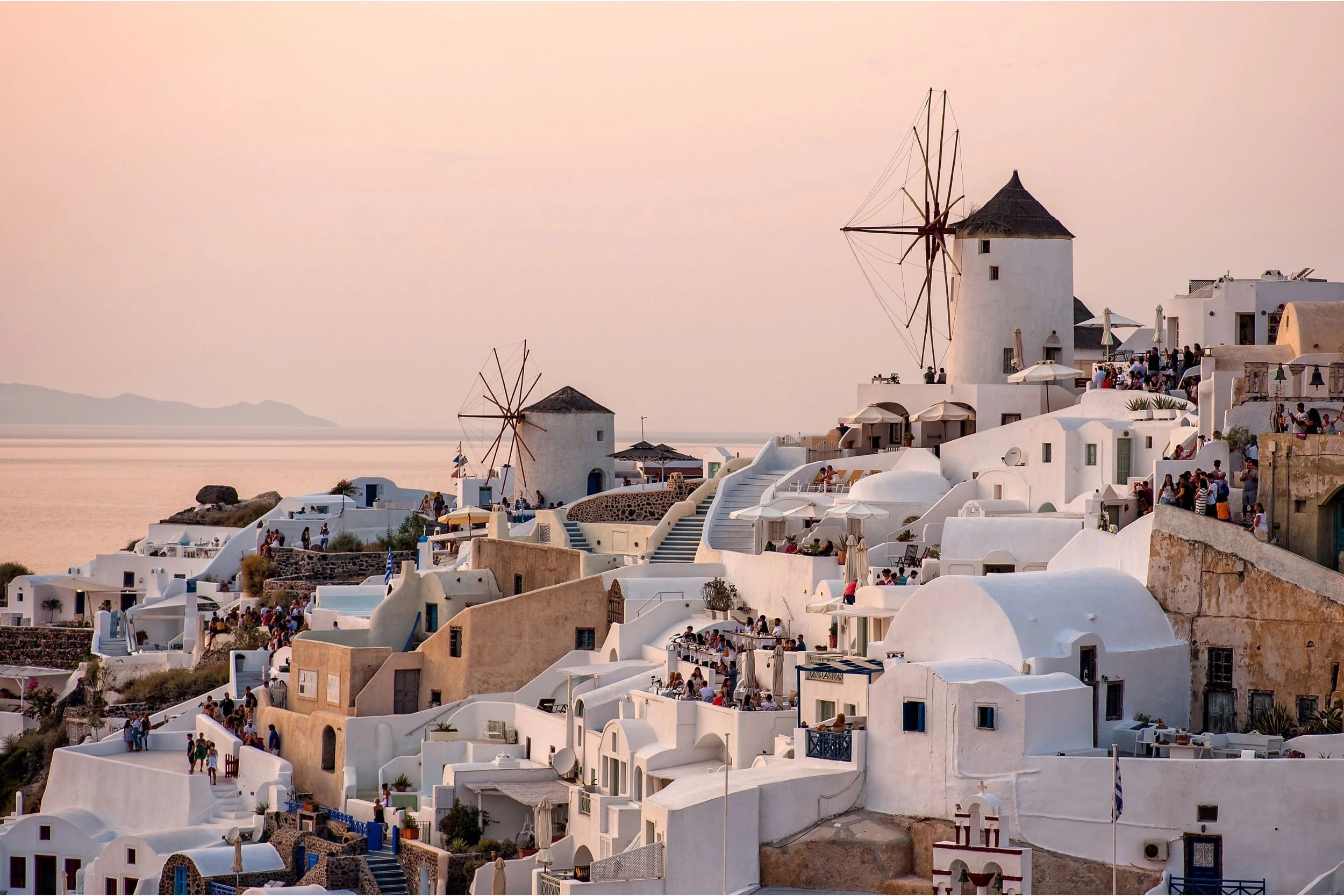 Traditional white-washed buildings with blue accents on a hillside in Santorini, Greece, with windmills and people enjoying the scene at sunset near the sea.