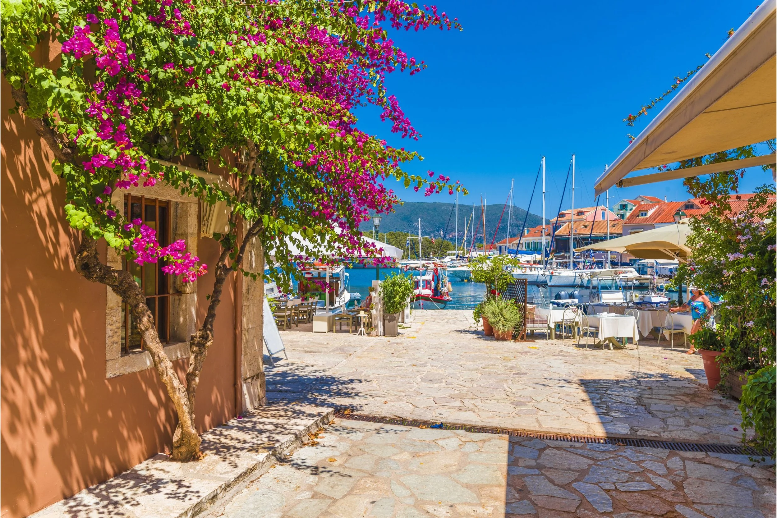 A scenic marina with sailboats docked by the waterfront, colorful buildings with red-tiled roofs, a stone-paved walkway shaded by a flowering tree with bright pink flowers, outdoor seating, and mountain in the background under a clear blue sky.