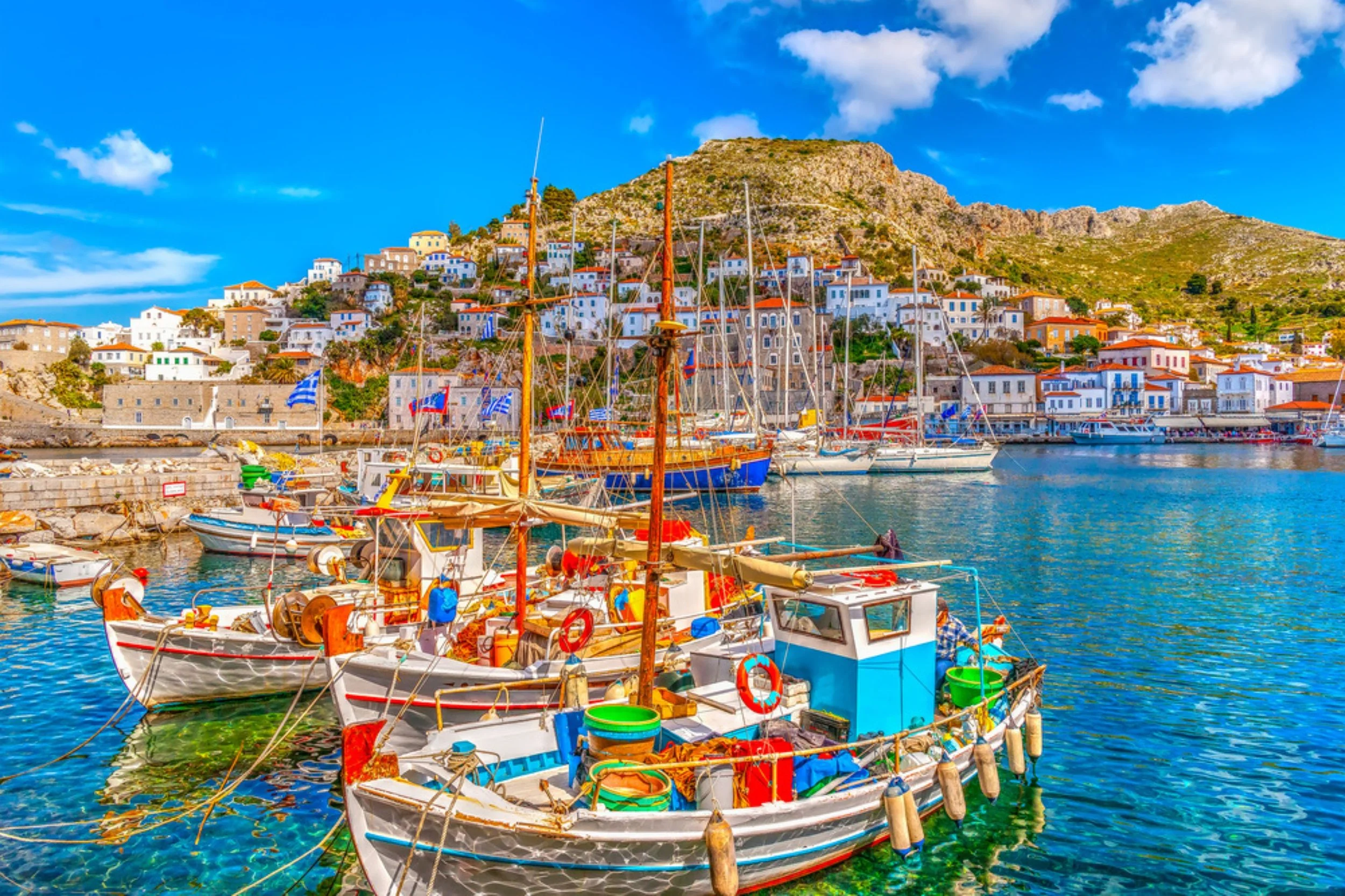 Colorful boats docked in a harbor with a hillside town and mountain in the background under a partly cloudy blue sky.