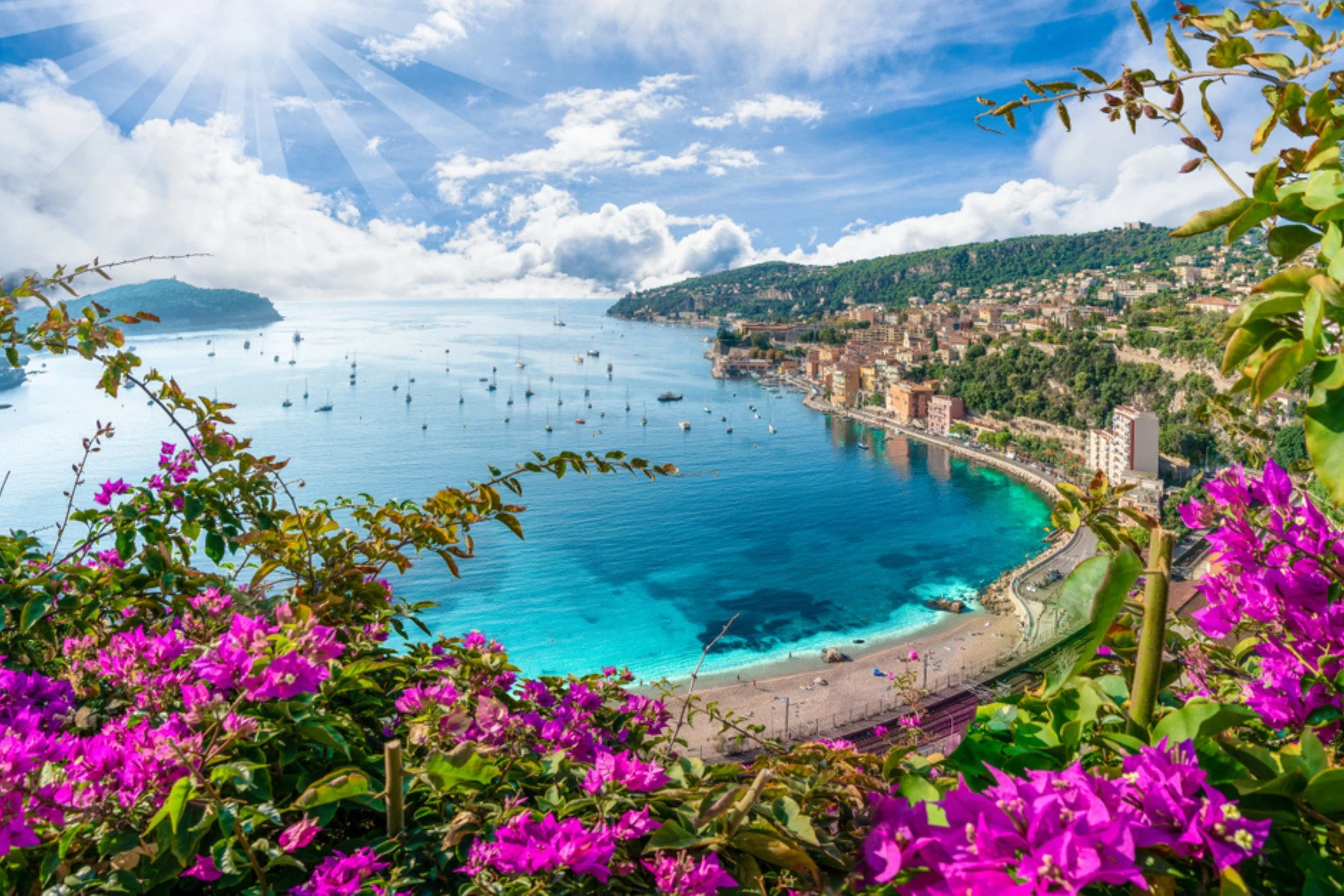 Coastal city with a crescent-shaped beach, turquoise water, boats in the harbor, hillside buildings, and pink flowering bushes in the foreground.