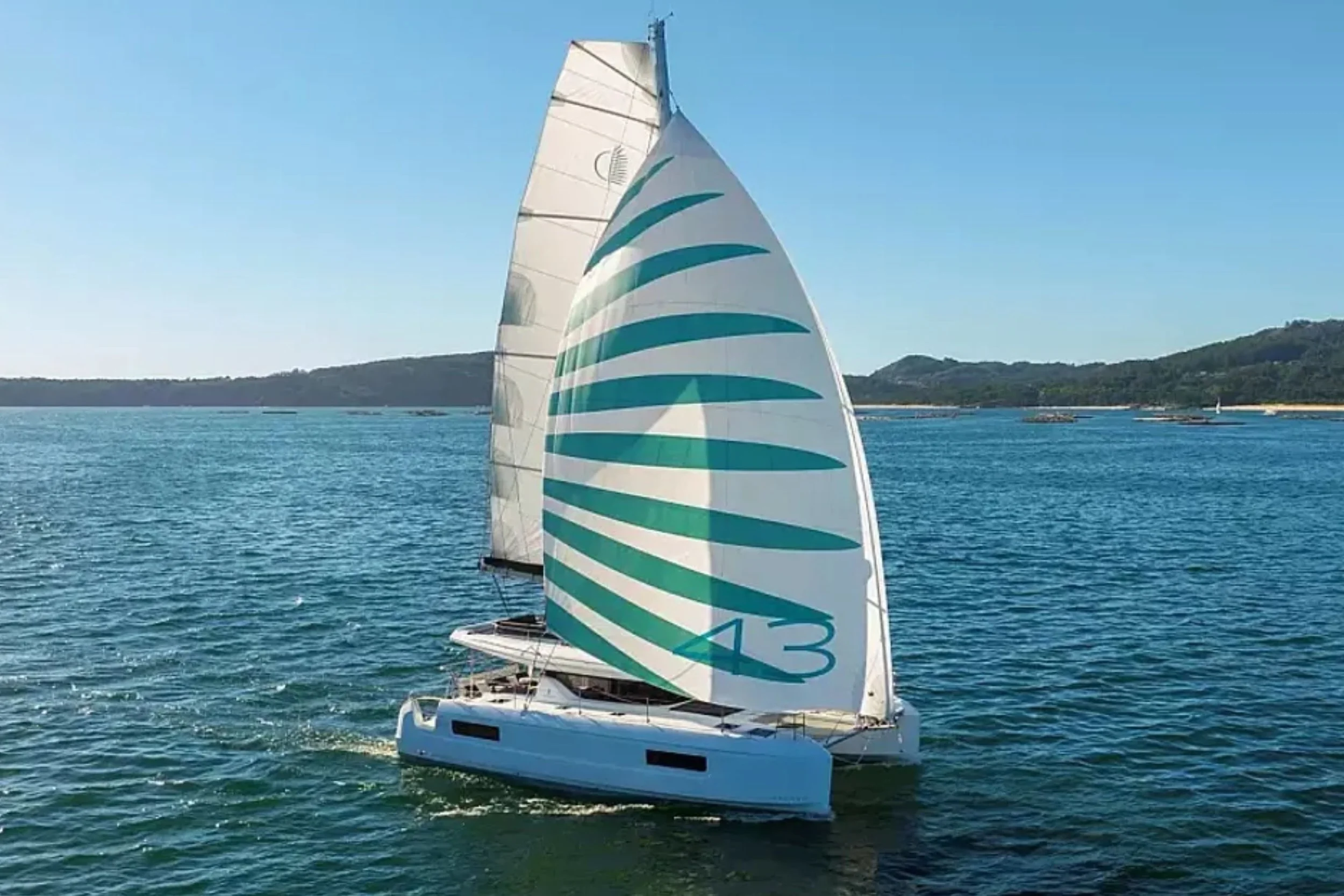 Sailboat with a distinctive, large sail marked with the number 43, sailing on a calm blue body of water under a clear sky, with land and hills in the background.