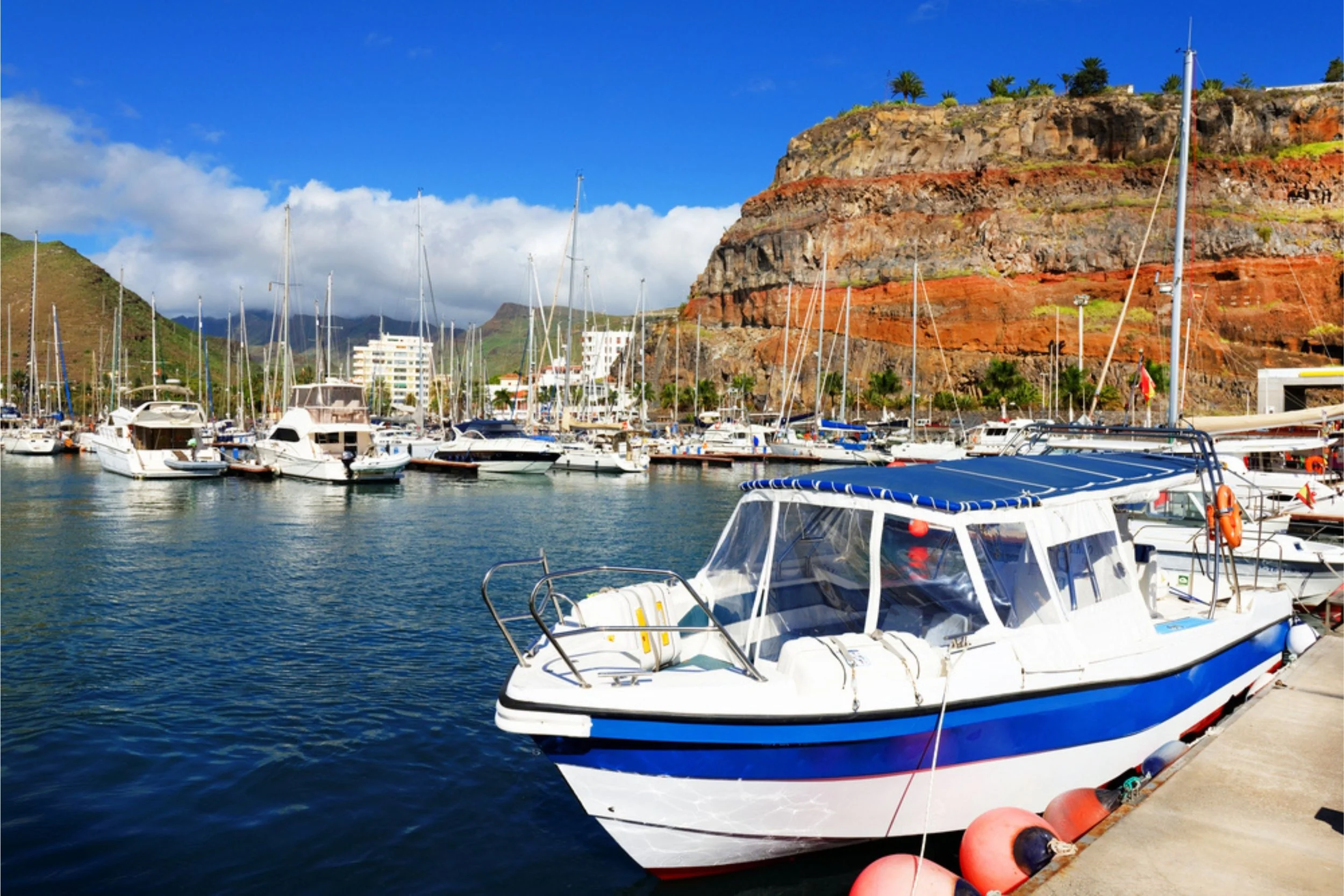 A marina filled with various boats and yachts docked by a concrete pier, with a red and brown rocky hillside and partly cloudy blue sky in the background.