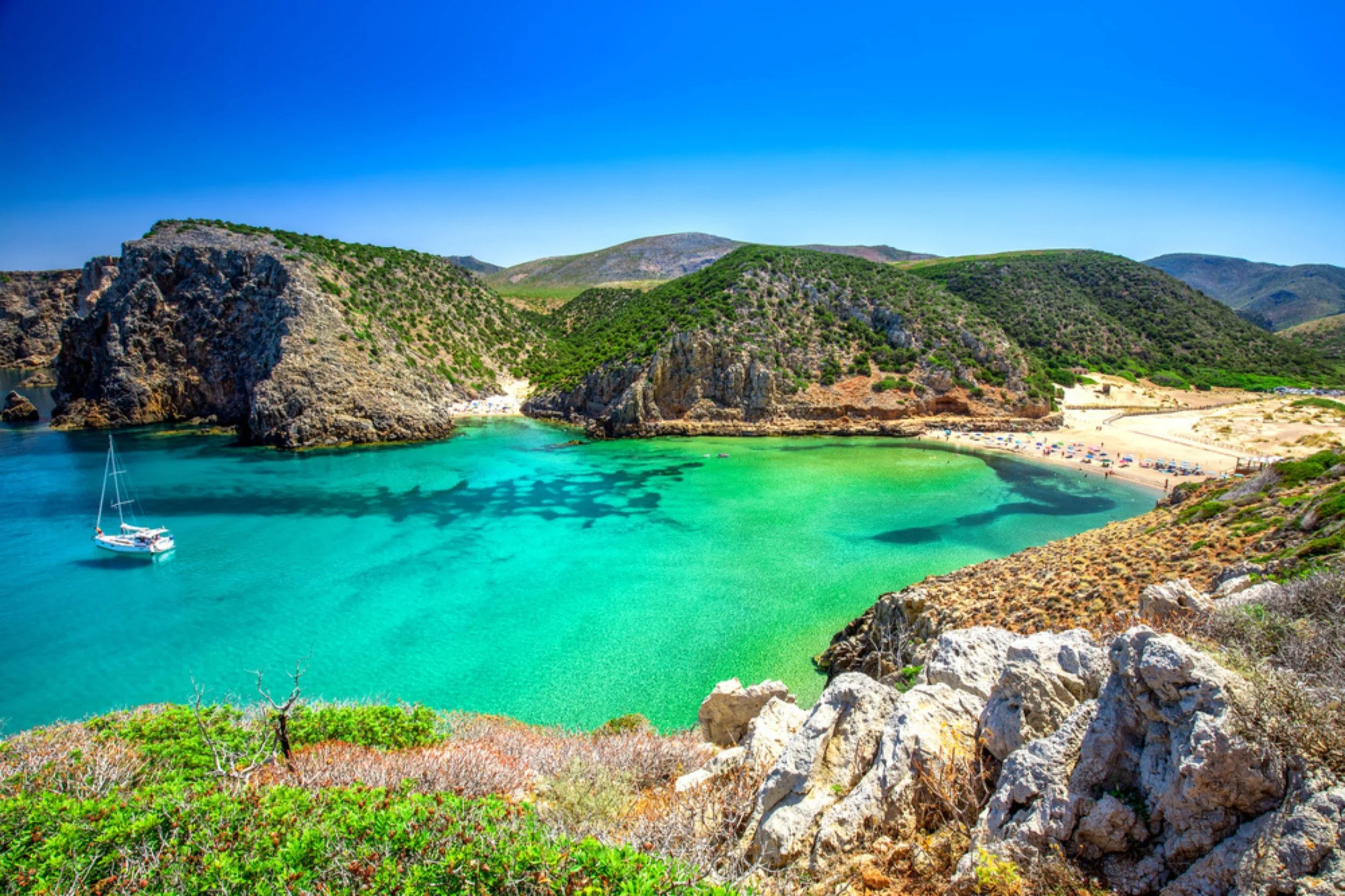 A scenic view of a cove with turquoise water, surrounded by rocky cliffs and green hills, featuring a sailboat anchored near the shore and a sandy beach with umbrellas.