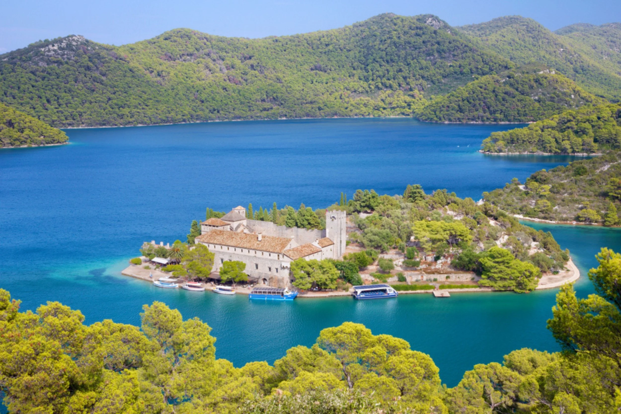 A scenic view of a small island with a historic stone building with a tower, surrounded by blue lake waters and lush green hills in the background.