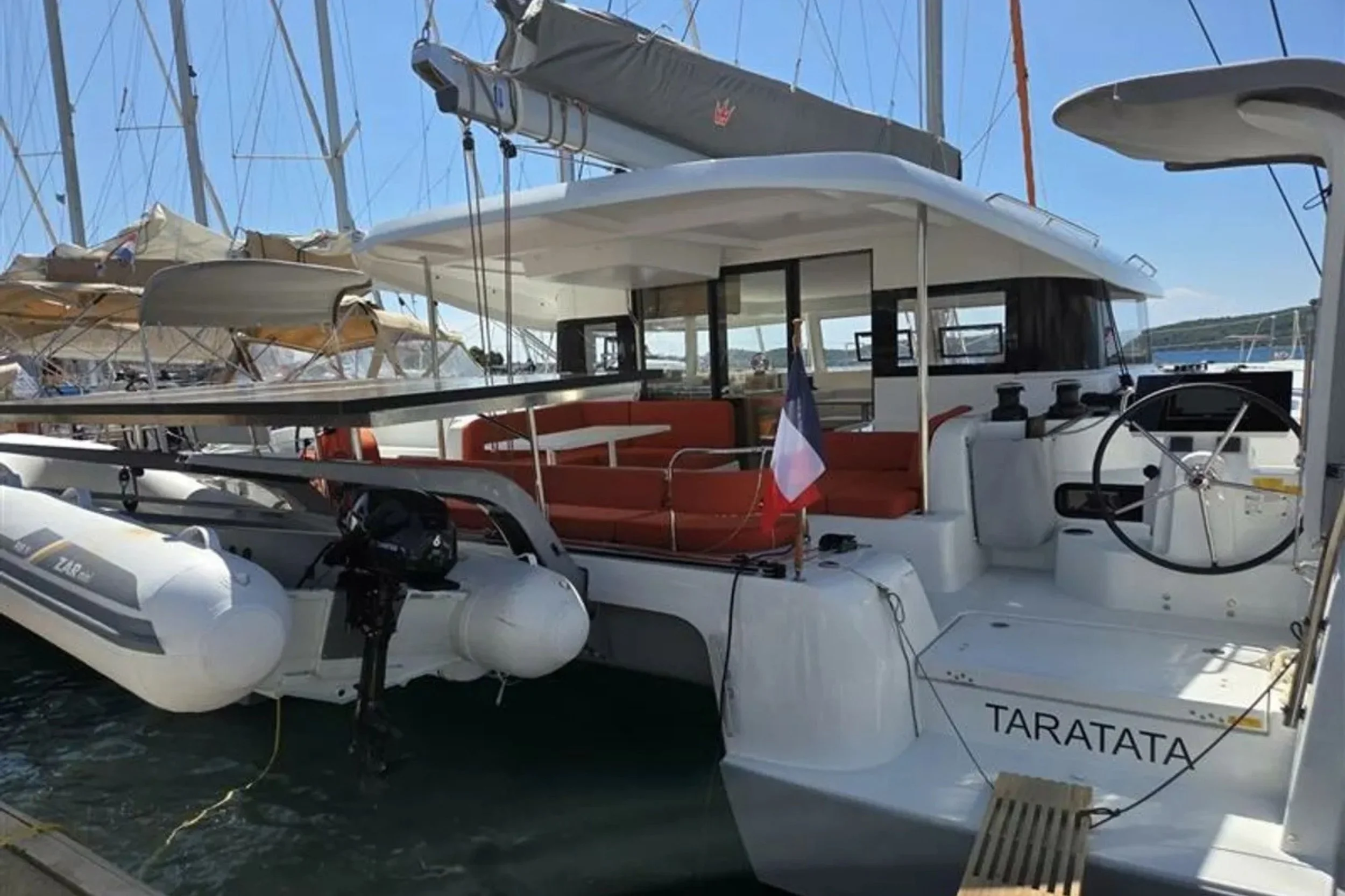 A white yacht named 'TARATATA' docked at a marina, with other sailboats in the background, featuring a small boat with a motor and a French flag on the rear deck.