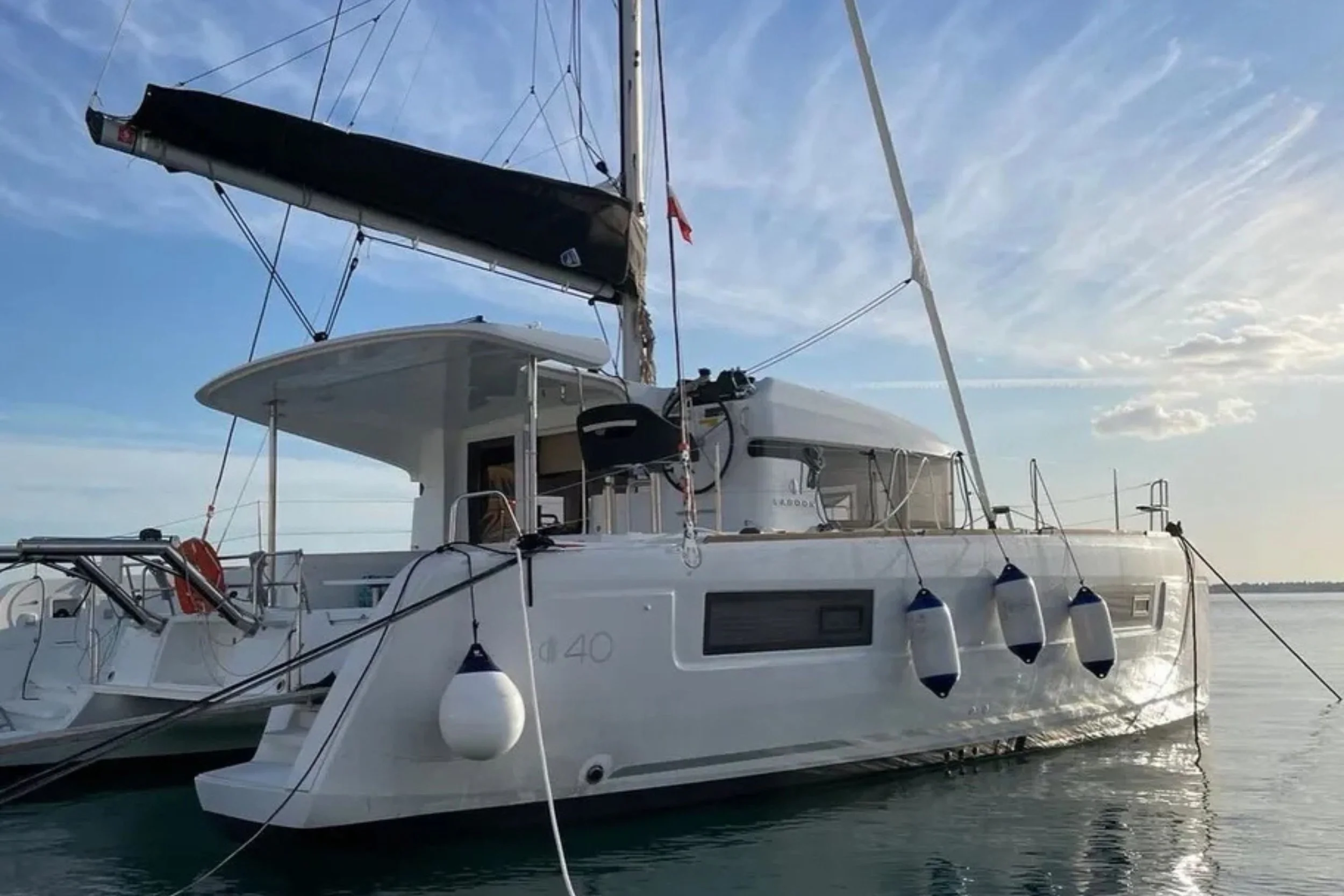 A white sailing yacht docked on calm water under a partly cloudy sky, with fenders hanging over the side and a black sail cover on the main sail.