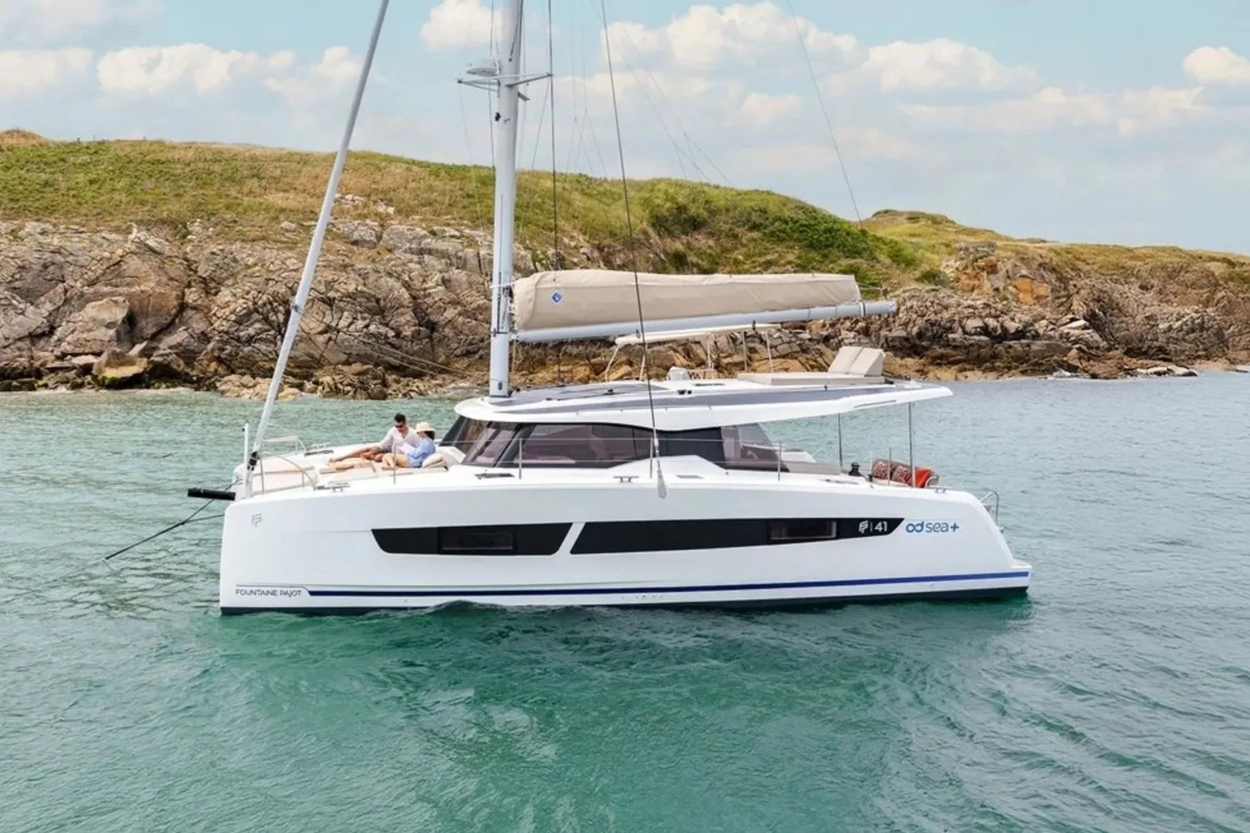 A white sailing yacht on turquoise water near rocky shoreline with grassy hills under a partly cloudy sky.