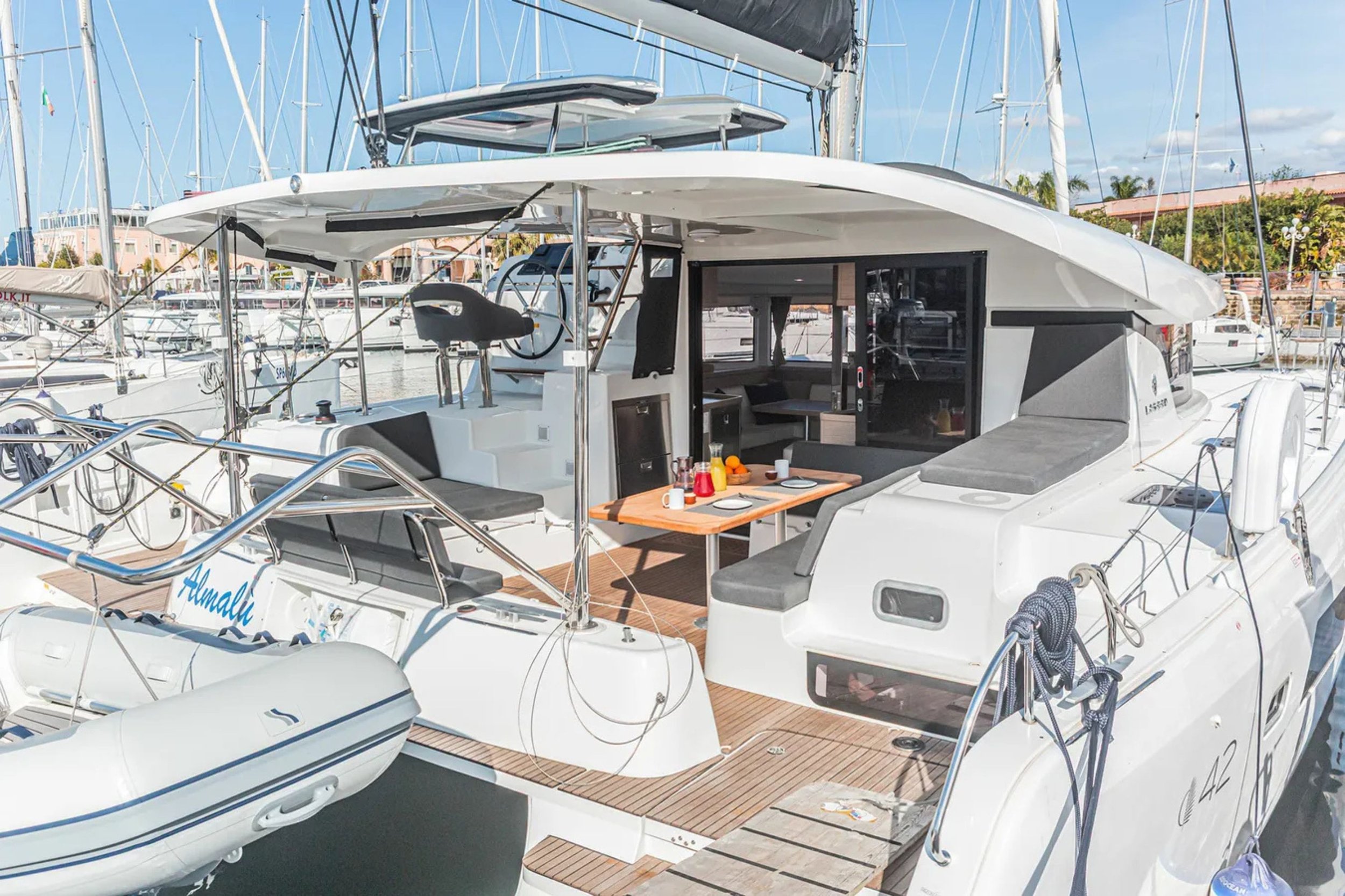 A white yacht docked at a marina, with outdoor seating, a dining table with breakfast items, and a view of other boats and sailboats in the background.