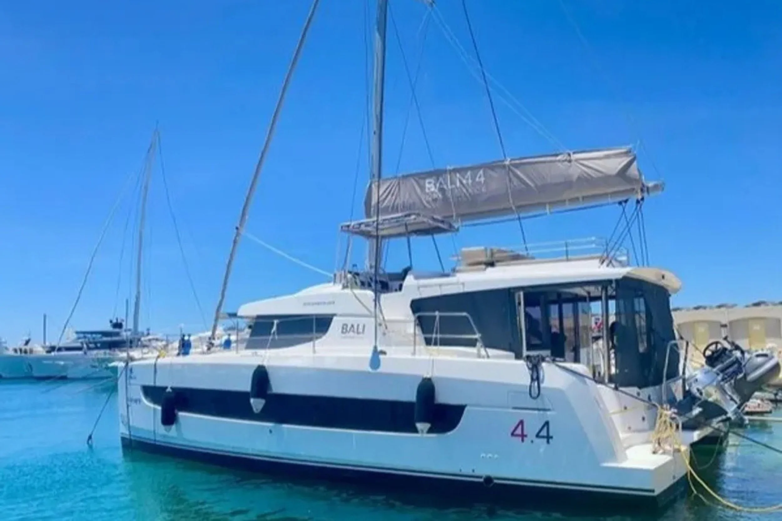 A white yacht named BALM with a black stripe, docked at a marina with clear blue sky and calm water.
