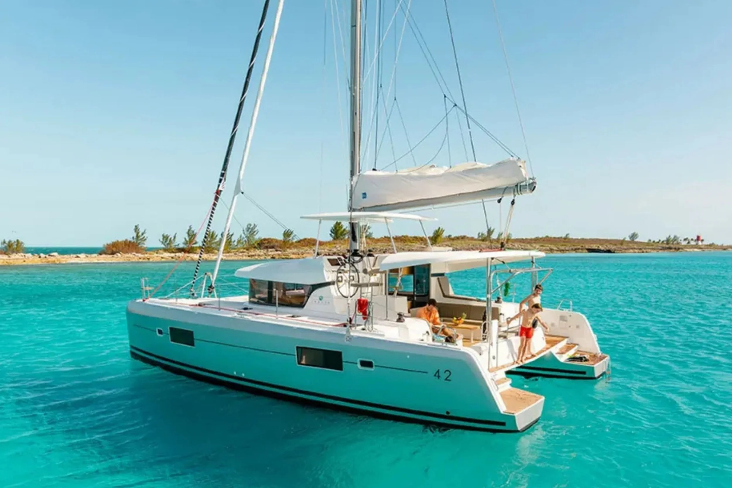A white sailboat with people on deck, anchored in clear blue water near a small island with sparse trees and a distant lighthouse under a clear sky.