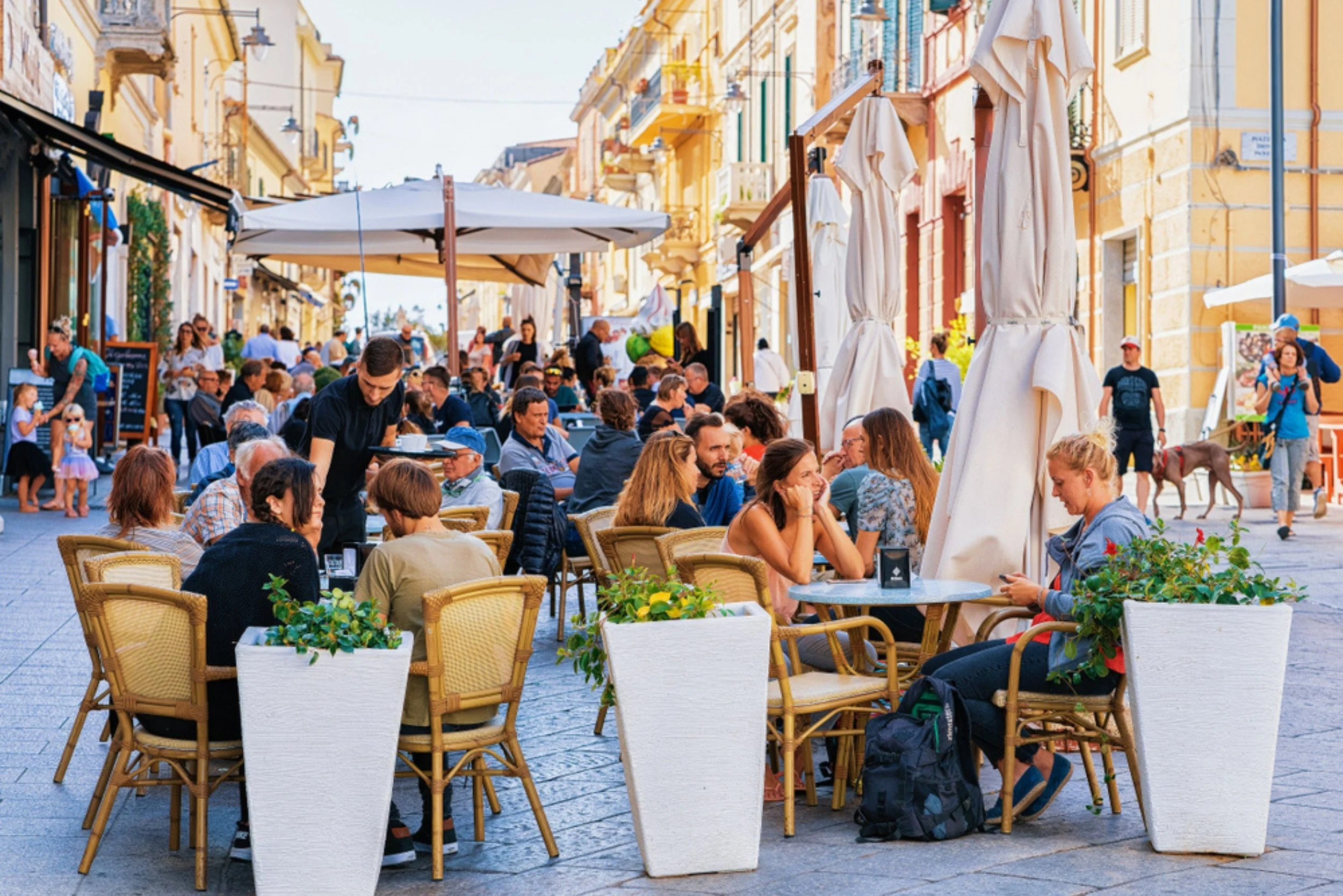Outdoor cafe filled with people dining, surrounded by potted plants, on a busy city street during daytime.