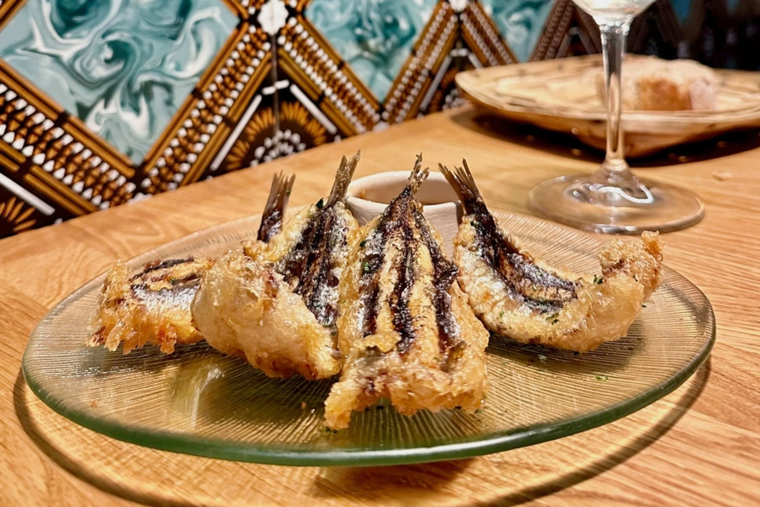 Fried fish on a glass plate, garnished with herbs, placed on a wooden table.
