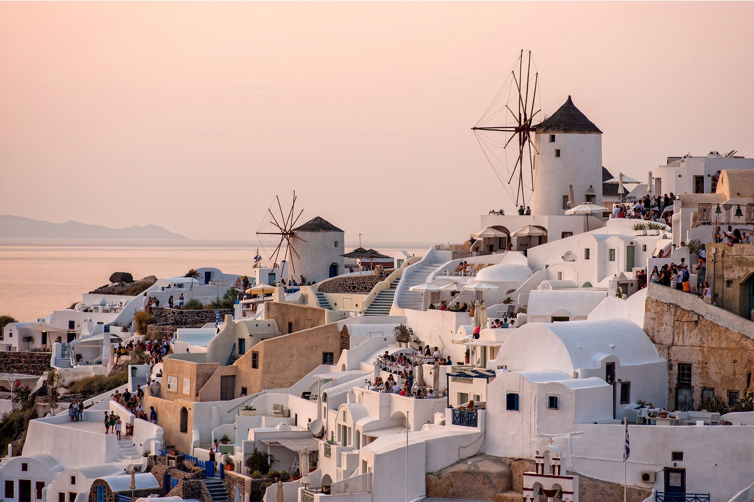 A picturesque coastal village in Santorini, Greece, with white buildings, windmills, terraces, and people enjoying the sunset view over the sea.
