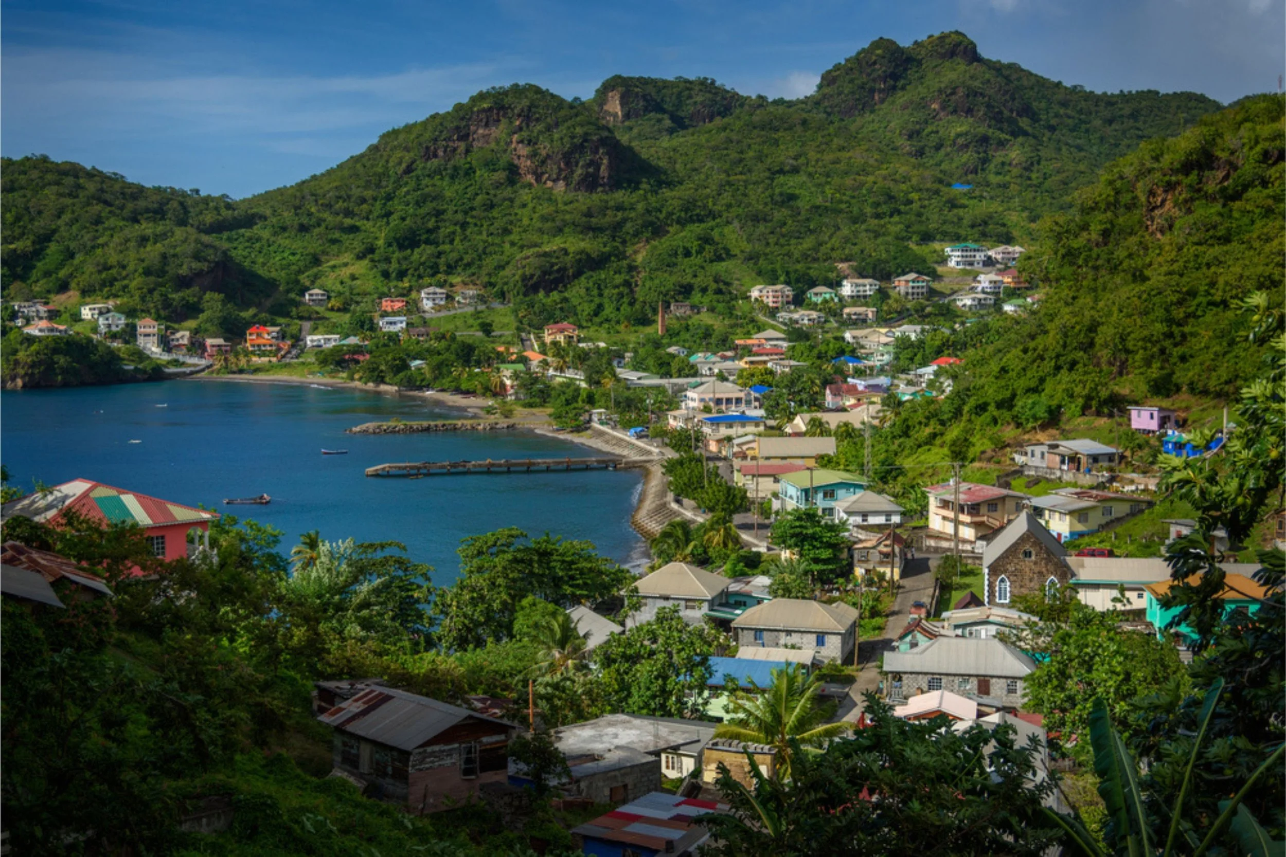Coastal village with colorful houses on a hillside, overlooking a bay with boats, surrounded by lush green mountains.
