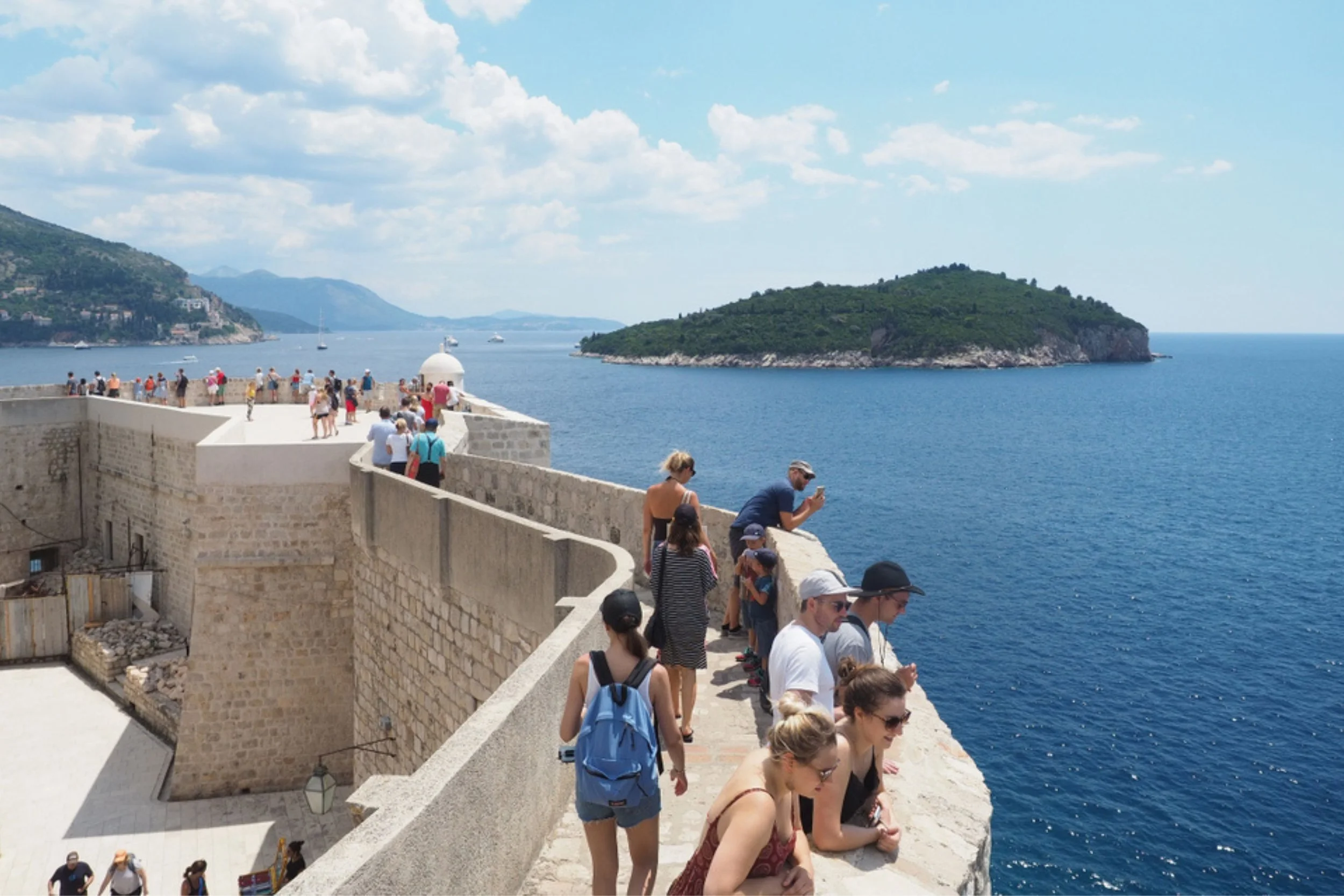 People walking and standing on a stone fortress wall overlooking the sea, with an island in the background on a sunny day with scattered clouds.