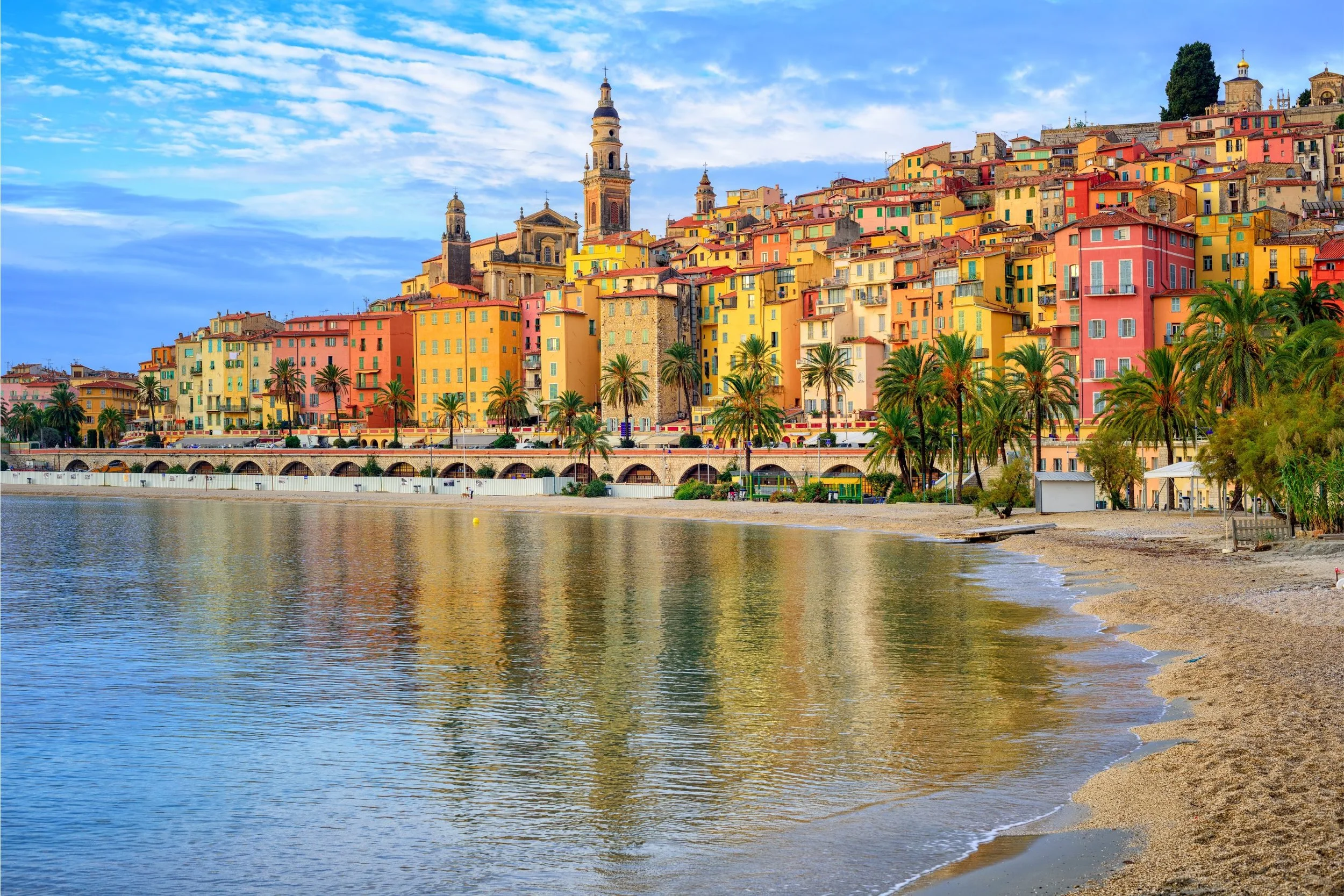 Colorful buildings and palm trees along a riverside in a Mediterranean town with a cloudy sky.