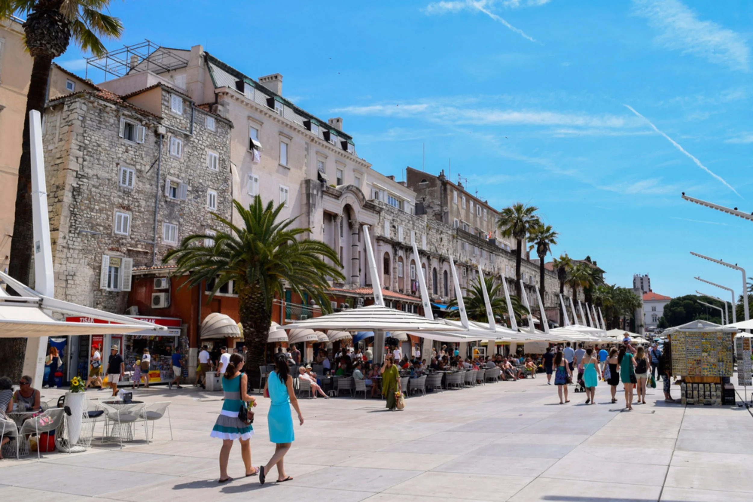 People walking and sitting at outdoor cafes along a promenade with palm trees, historic stone and stucco buildings, and a bright blue sky.