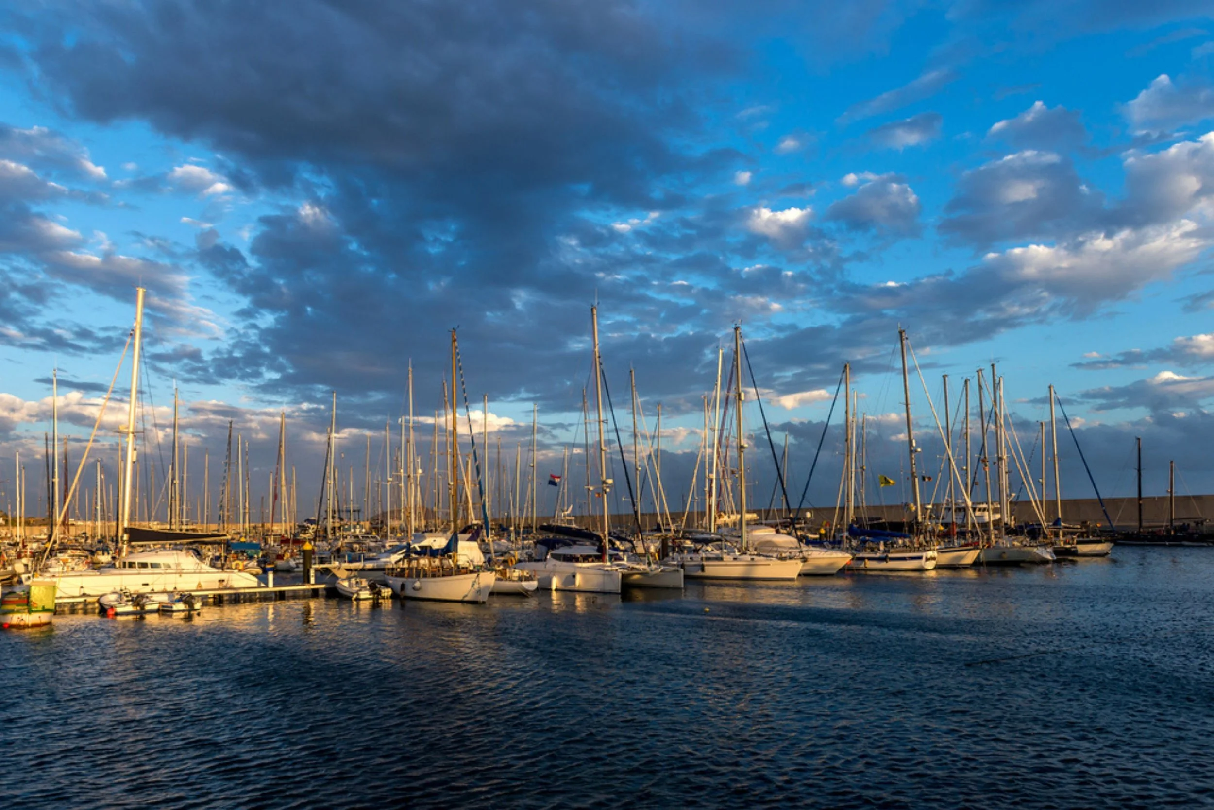 Several sailboats docked at a marina during sunset with a partly cloudy sky reflected on the water.