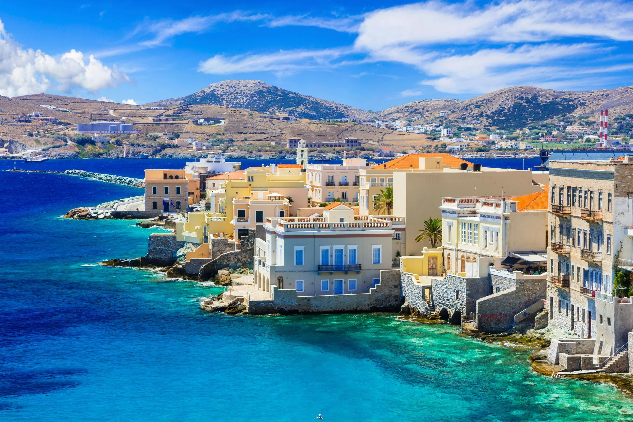 Colorful seaside town with buildings lining the rocky coast, turquoise water, mountain hills in the background, and a blue sky with clouds.