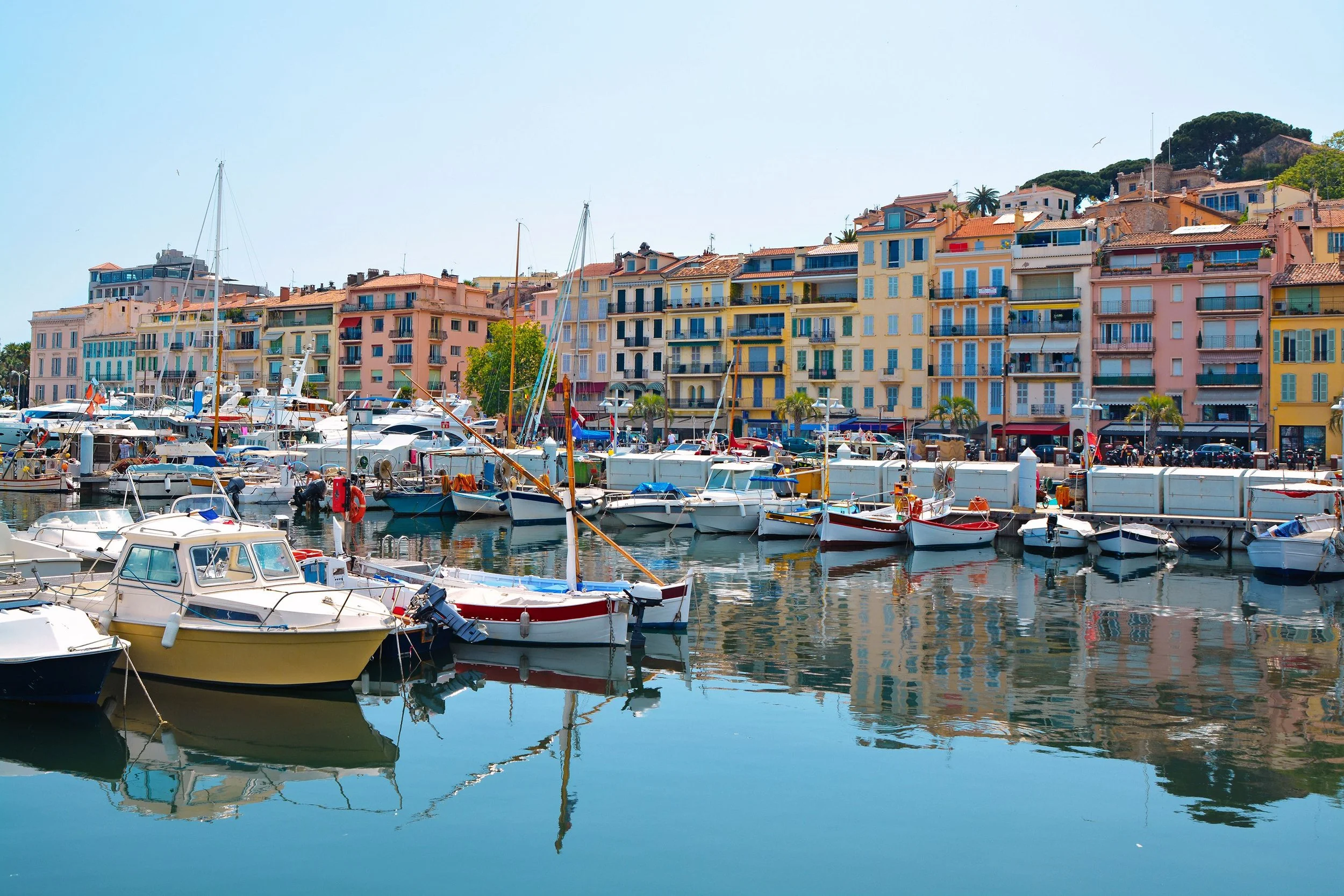 Colorful Mediterranean-style buildings along a marina filled with various sailboats and yachts, with calm water reflecting the boats and buildings.