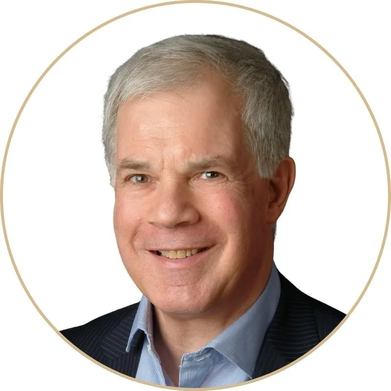 A headshot of a middle-aged man with gray hair, smiling, wearing a suit jacket and a light blue collared shirt, set against a white background with a thin gold border.