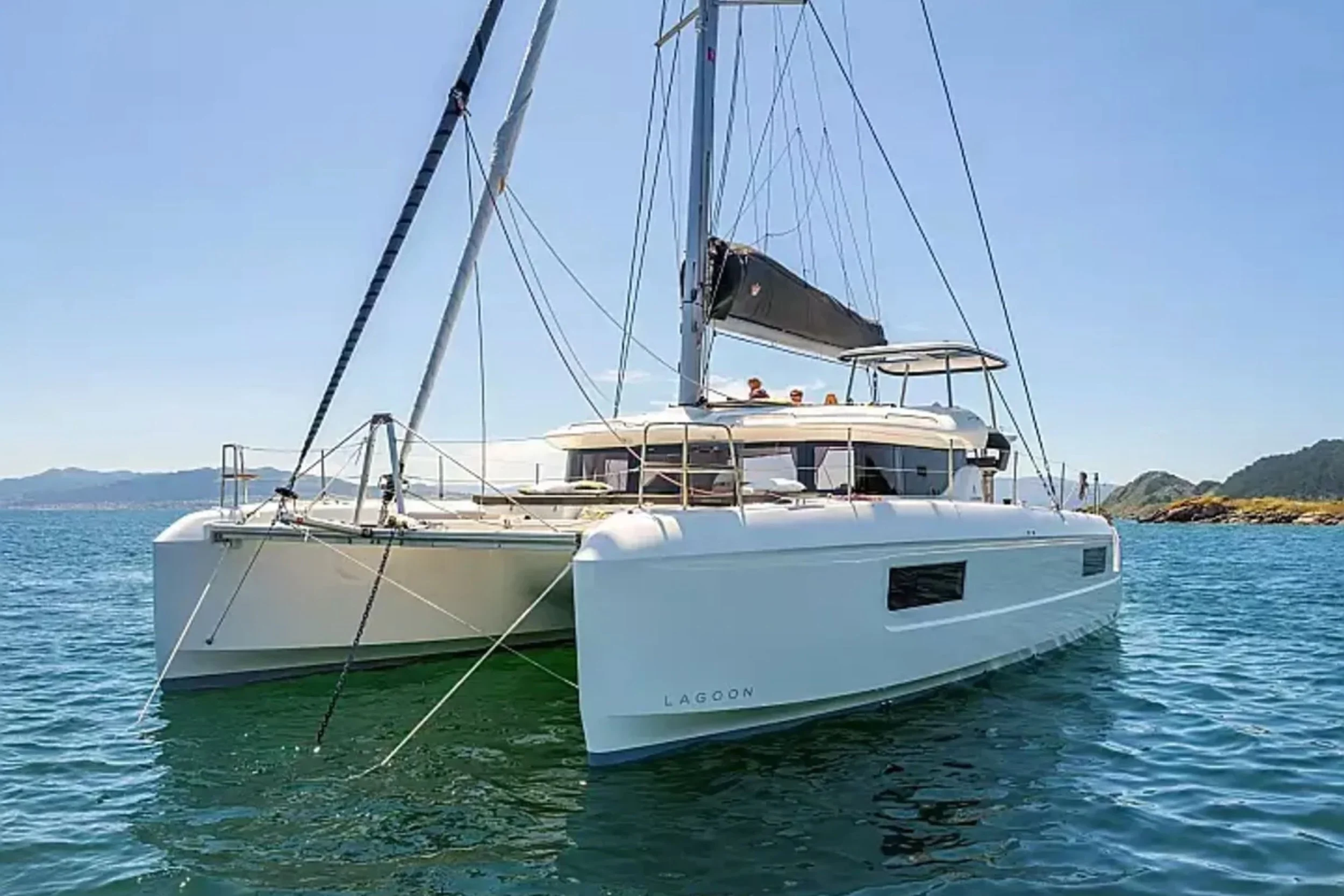 A white Lagoon sailing catamaran with a black sail, anchored in calm blue water with a distant shoreline and mountains in the background. There are people on the deck.