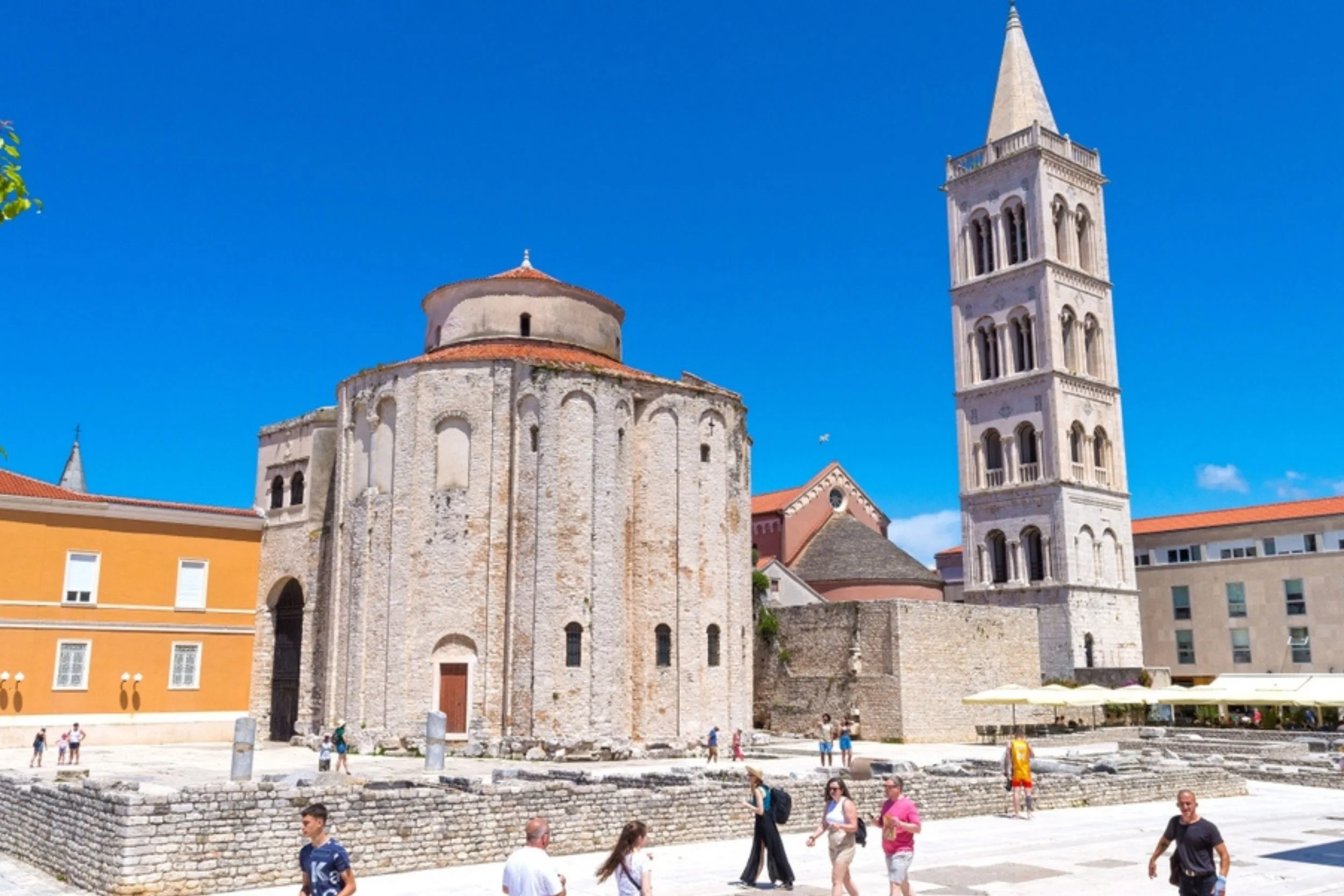 Tourists walking near ancient stone building with a church and bell tower under a clear blue sky.