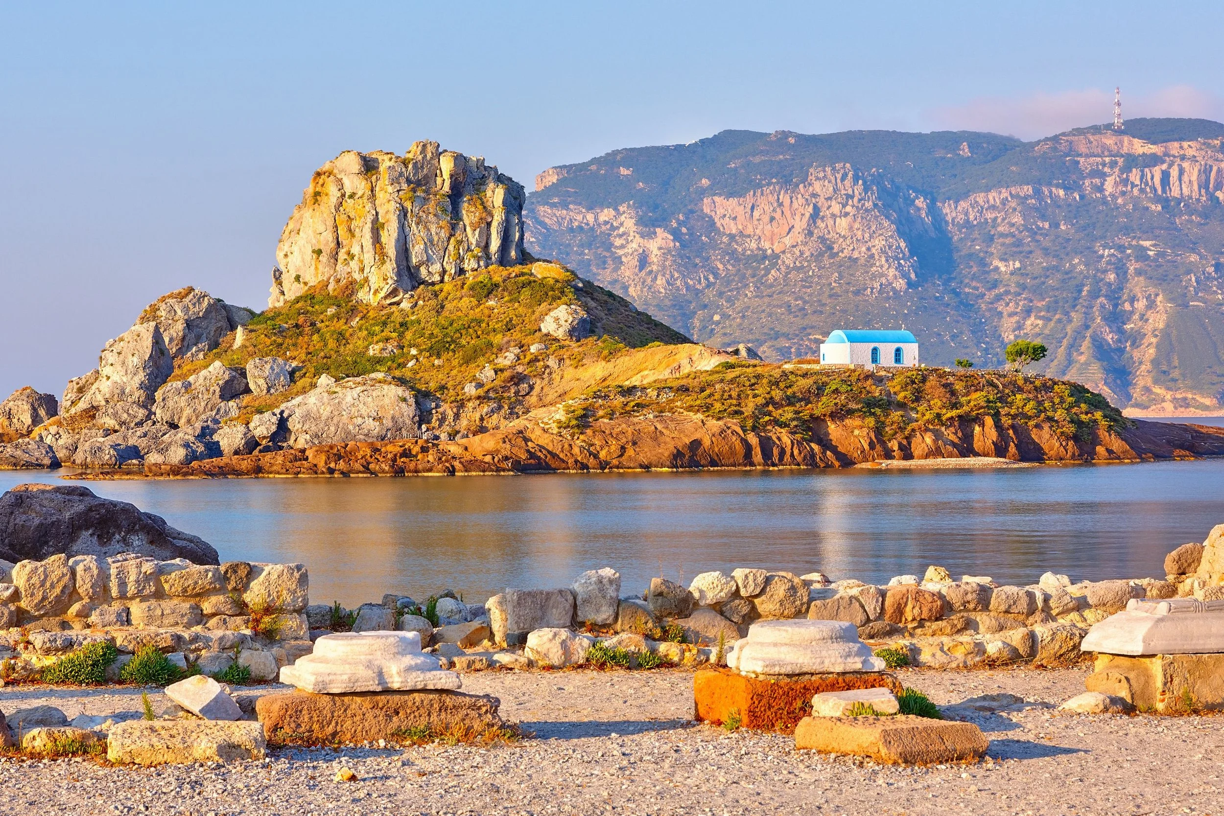 A coastal landscape with a rocky hill, calm water, and a small white house with a blue roof in the distance, under a light sky.