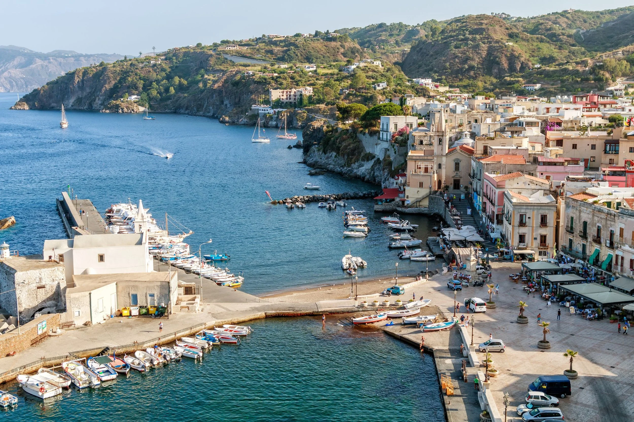 View of a seaside marina with boats docked, surrounded by colorful buildings on a hillside, in a coastal town.