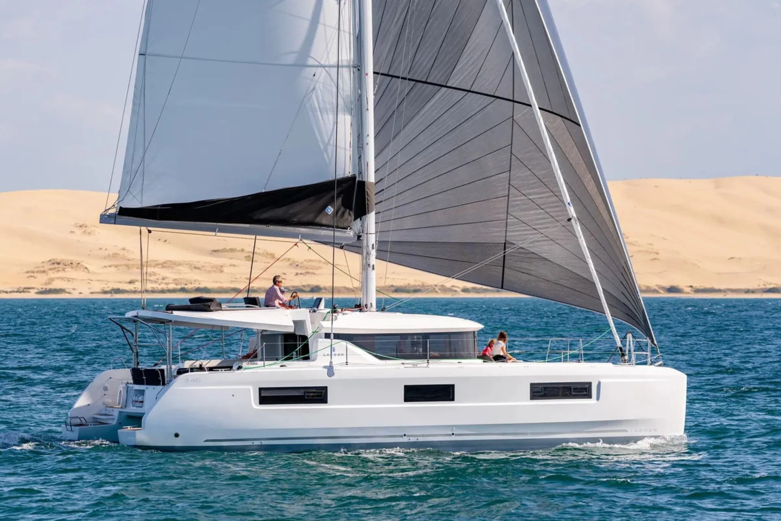 A white sailing catamaran cruising on blue water with a desert landscape in the background, featuring two people on deck and large sails.