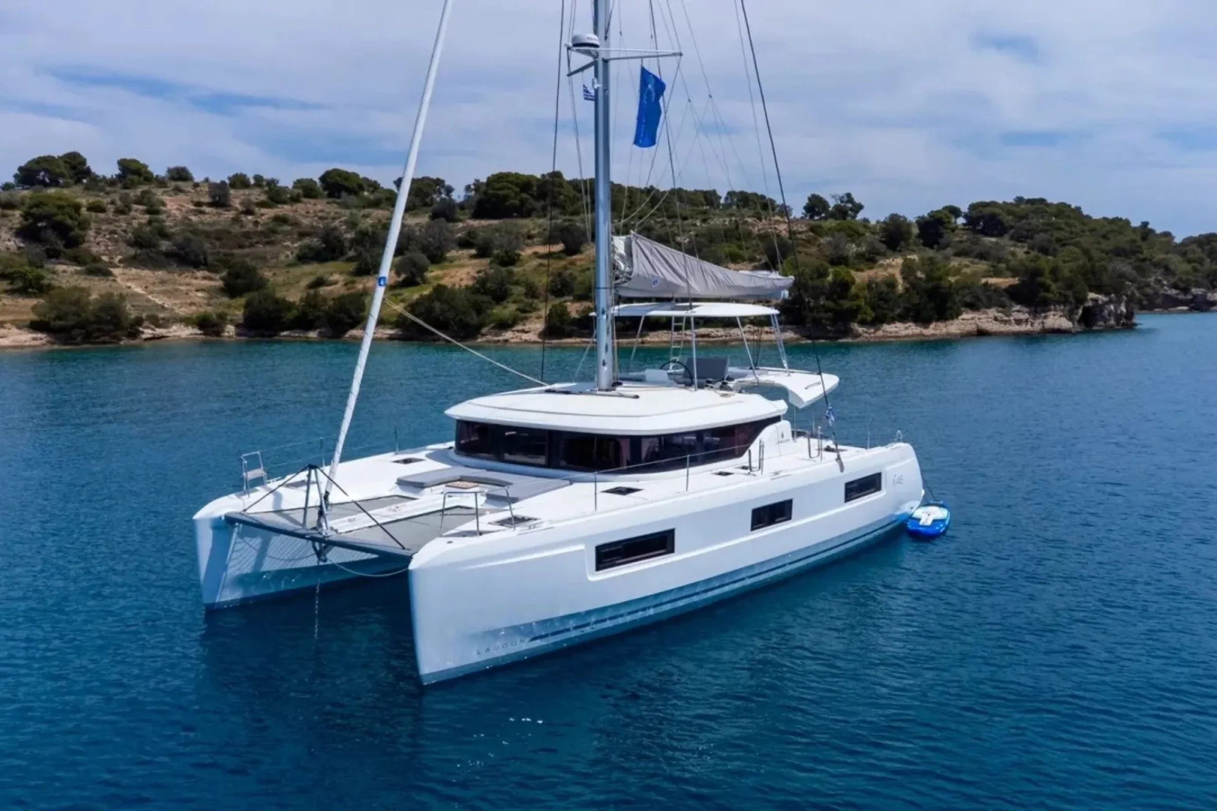 A white sailing catamaran anchored in clear blue water near a hilly, shrub-covered coastline.