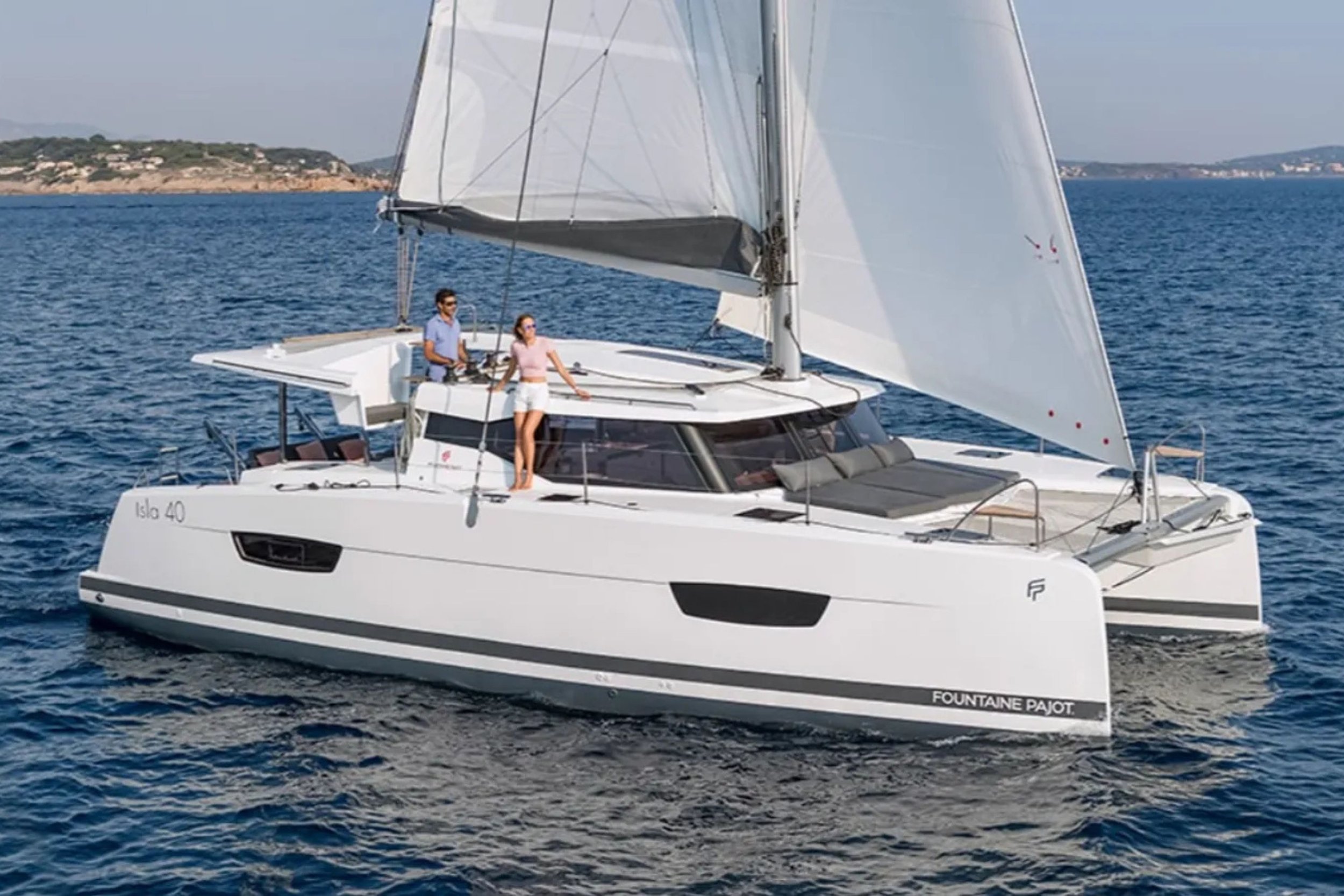 A white Fountain Pajot catamaran sailing on the ocean with two people on board, a man and a woman, enjoying the view. The boat has large sails and a sleek modern design, with a distant shoreline visible in the background.