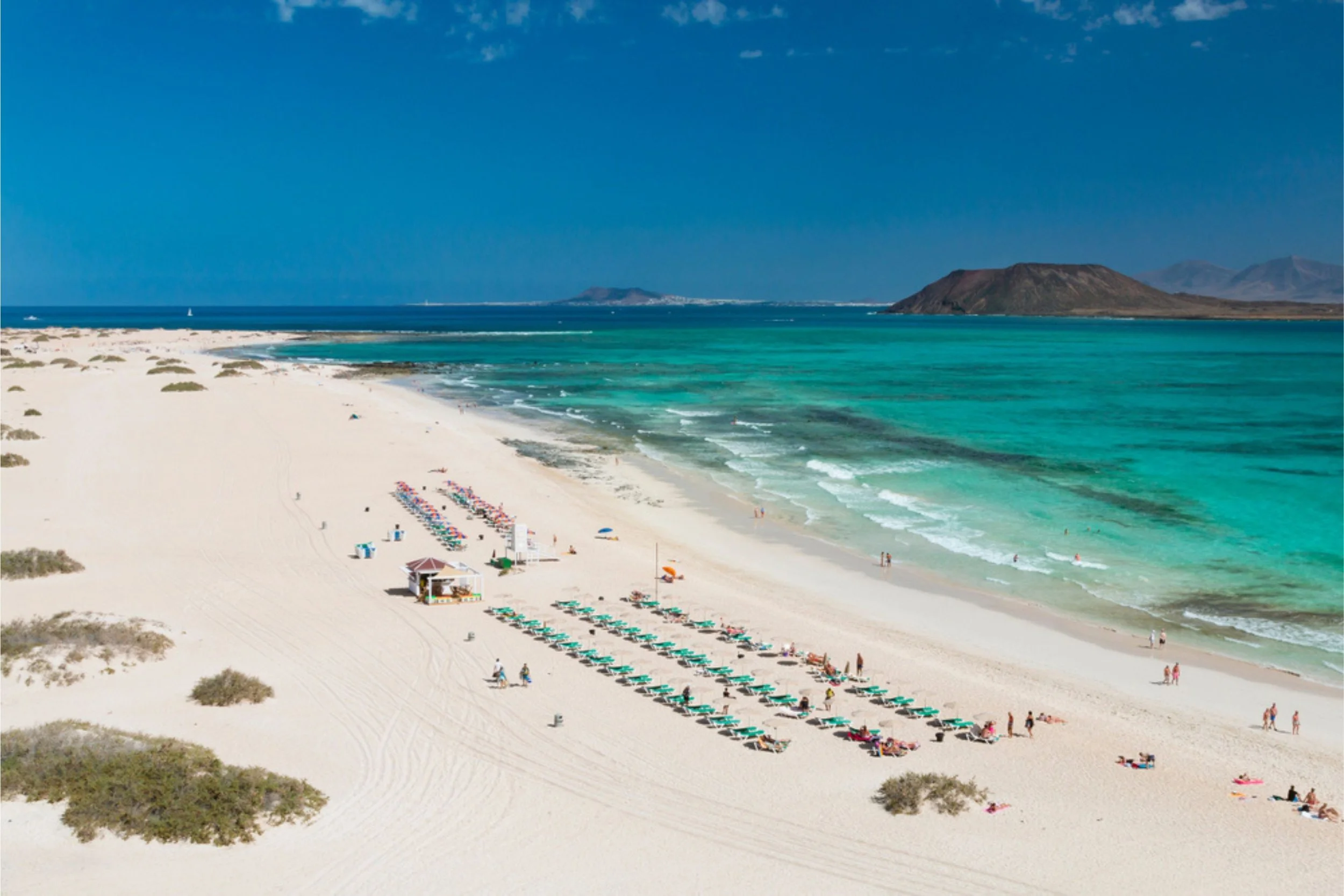 Bird's eye view of a beach with white sand, turquoise water, and mountains in the background. There are rows of lounge chairs and umbrellas, some small structures, and people relaxing and walking along the shoreline.