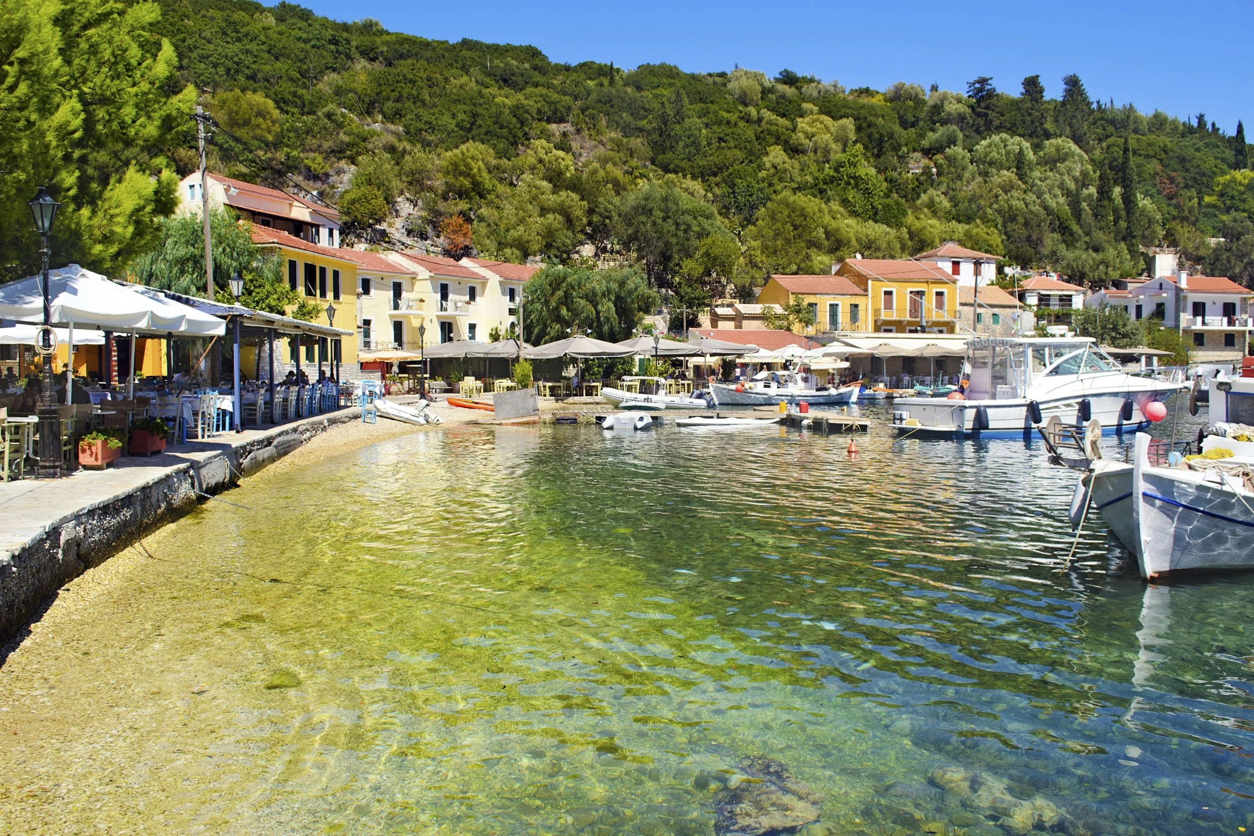 A small harbor with boats docked along the shoreline, colorful houses in the background, and outdoor seating with umbrellas on the left side, surrounded by lush green hills.