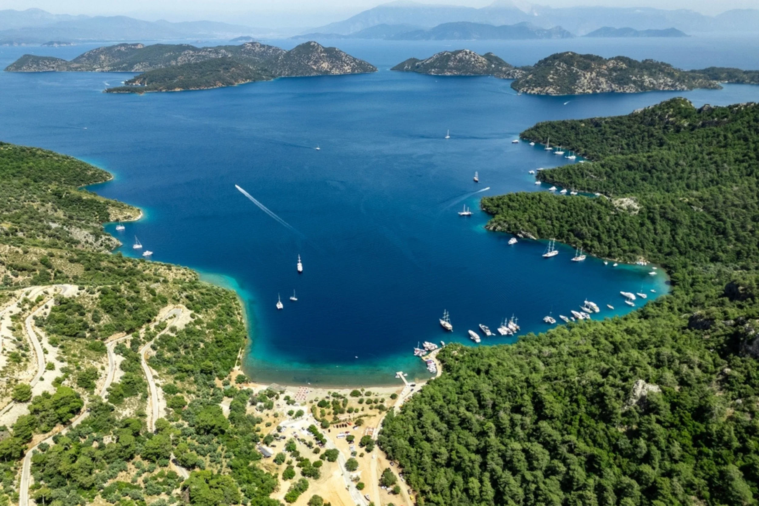 An aerial view of a bay with boats and sailboats, surrounded by lush green hills and rocky islands in the distance.