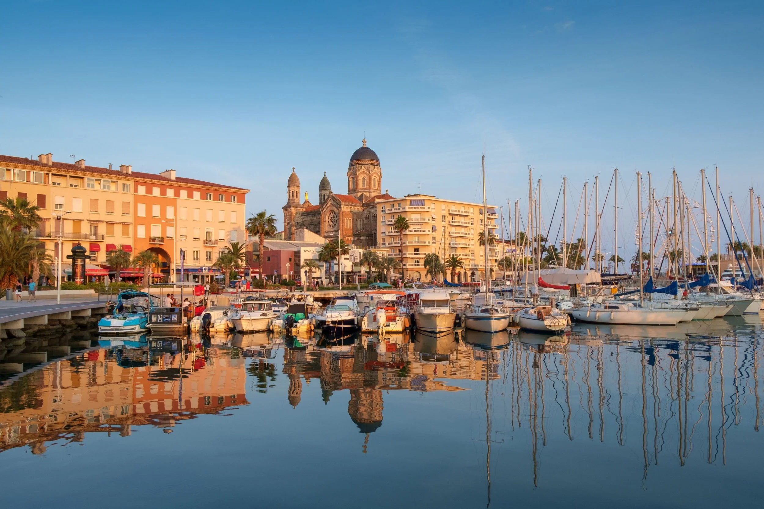 Boats docked at a marina with colorful buildings and a church in the background, under a clear blue sky.