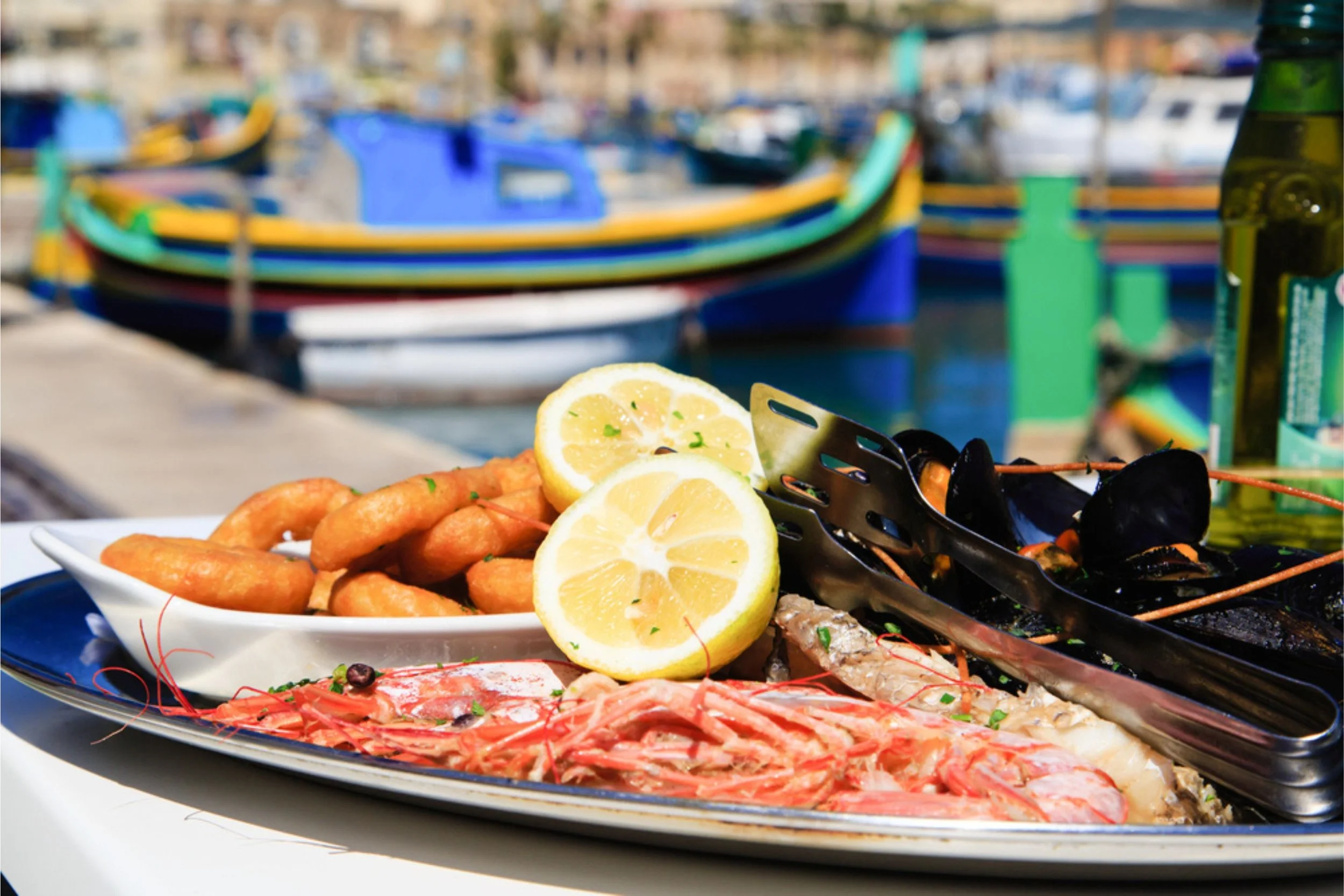 Seafood platter with lemon slices, fried fish, shrimp, and mussels near a harbor with boats.