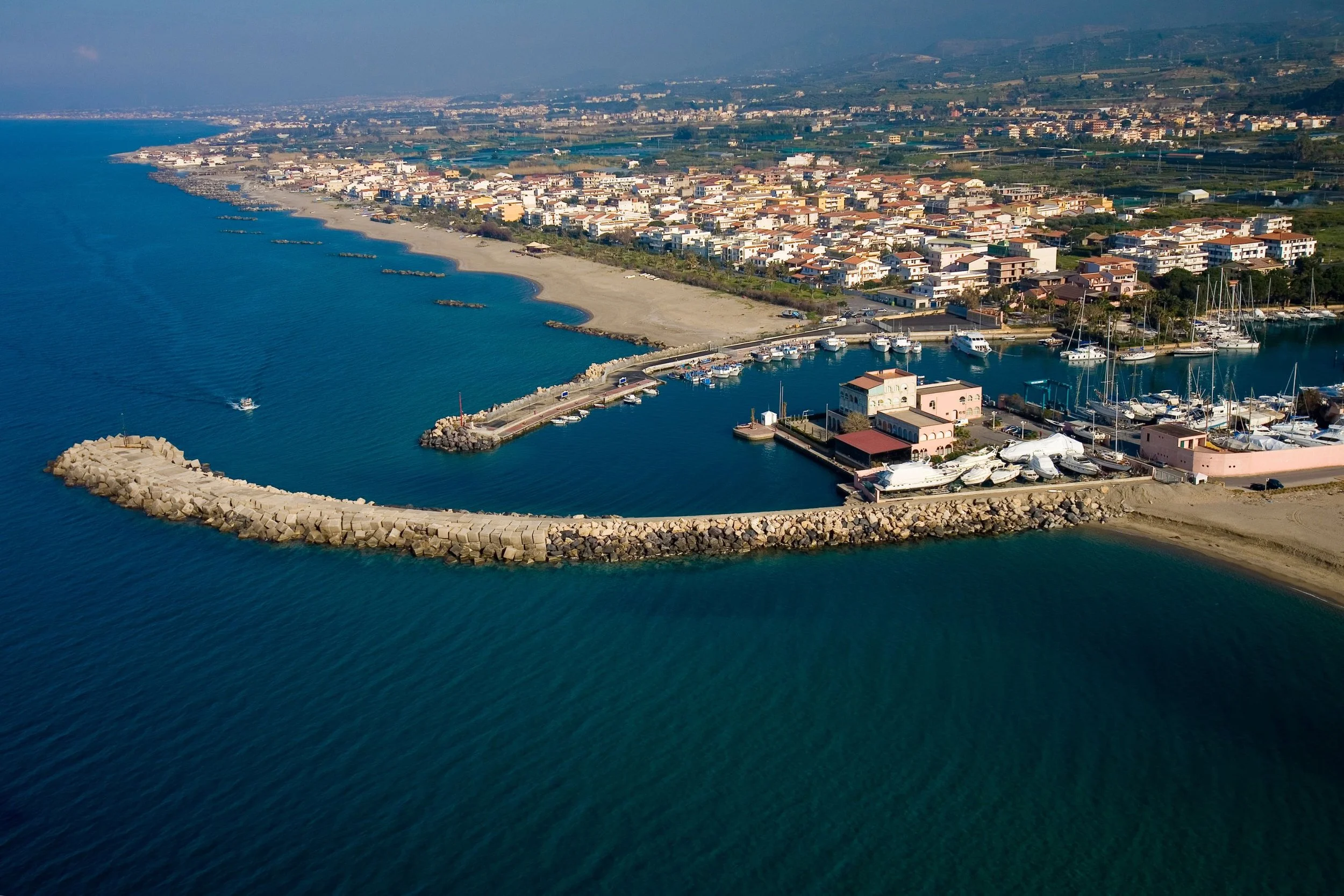 Aerial view of a marina with boats docked, surrounded by a breakwater, and a coastal town with buildings and a beach, extending into a bay with calm blue water.