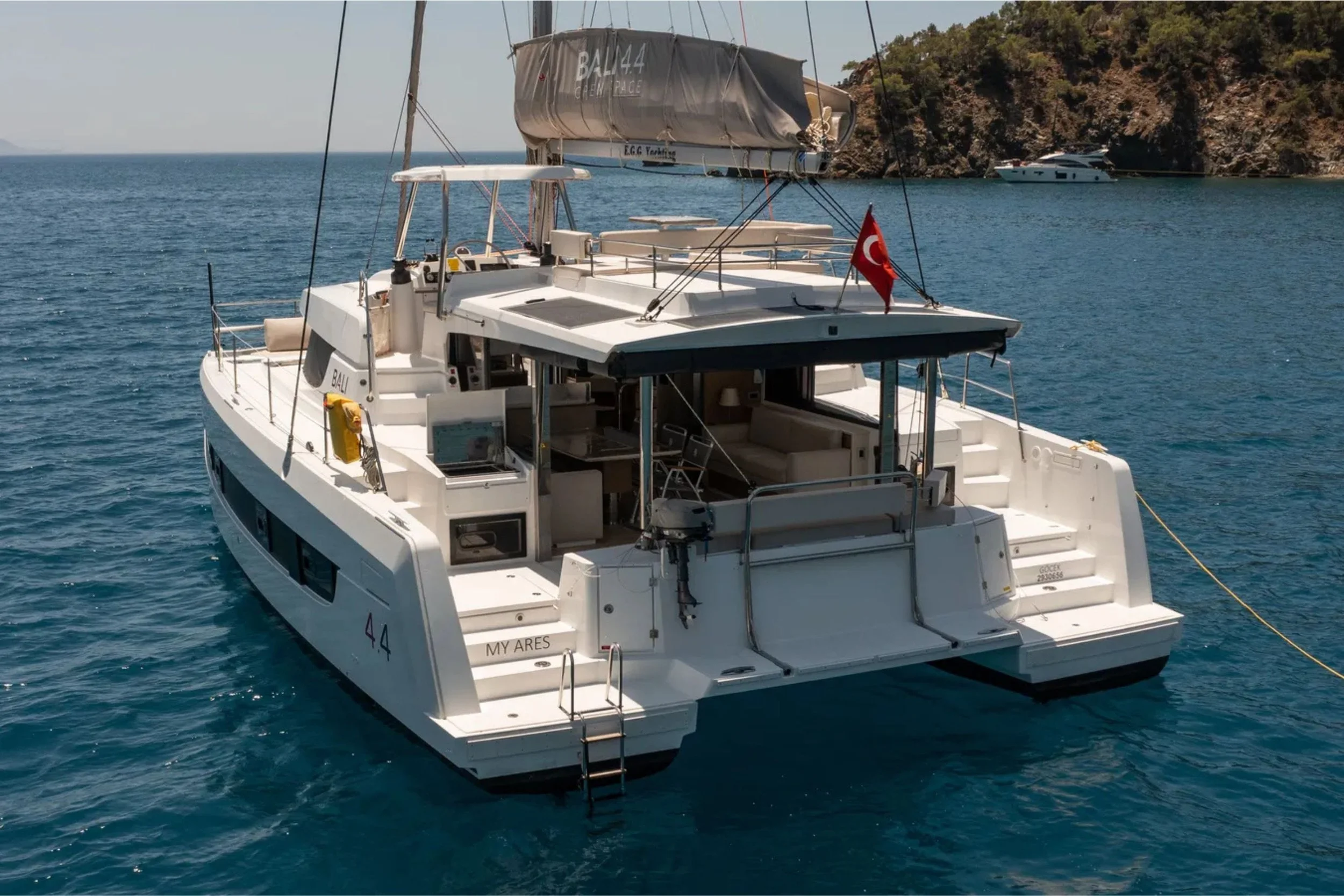 A white luxury yacht named MY ARES anchored in blue water near a rocky hillside, with Turkish flag on the stern and an additional large yacht in the background.