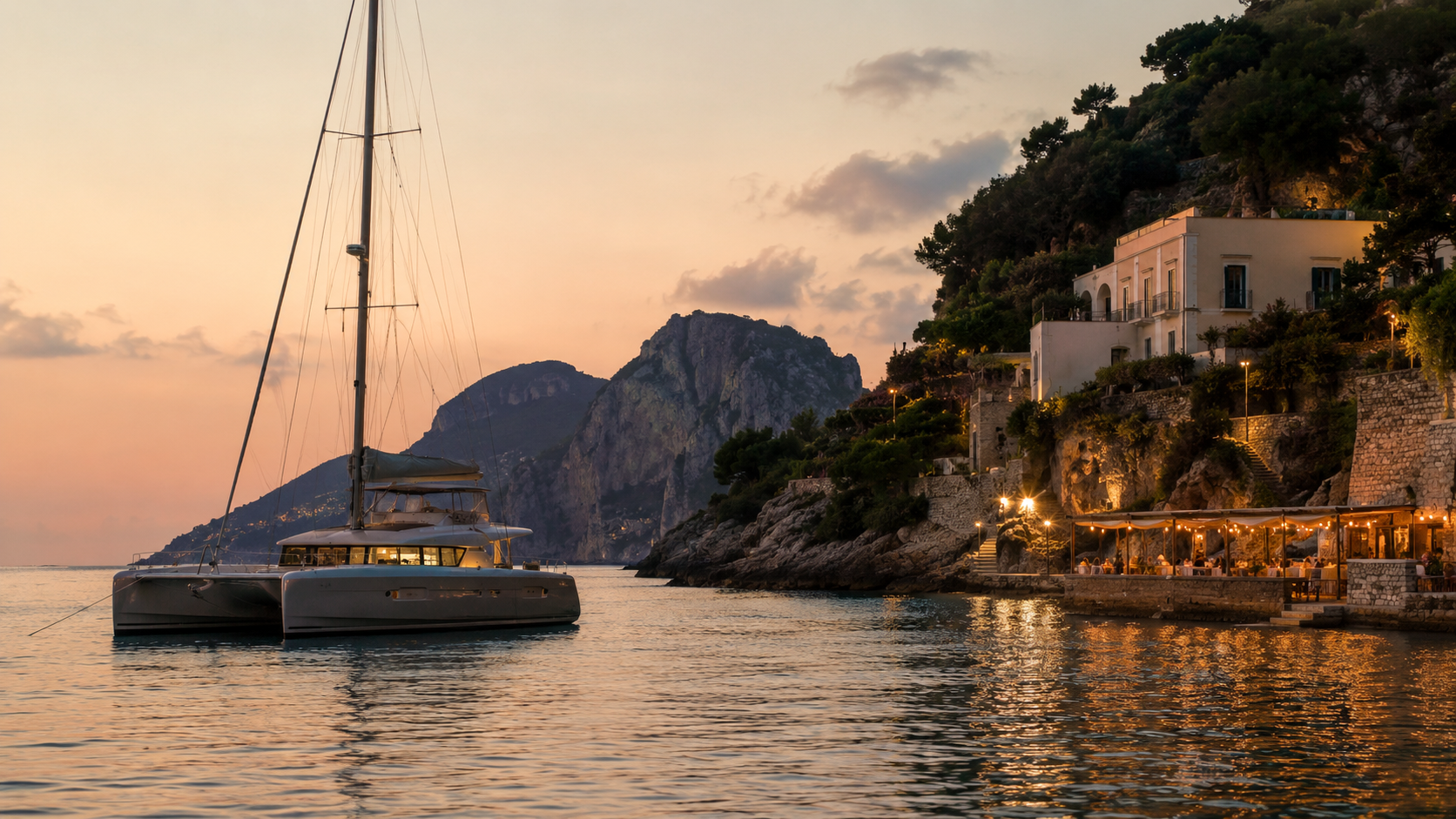 Modern catamaran anchored in a peaceful Italian coastal harbour at dusk, with warm waterfront restaurant lights and rocky island hills beyond.