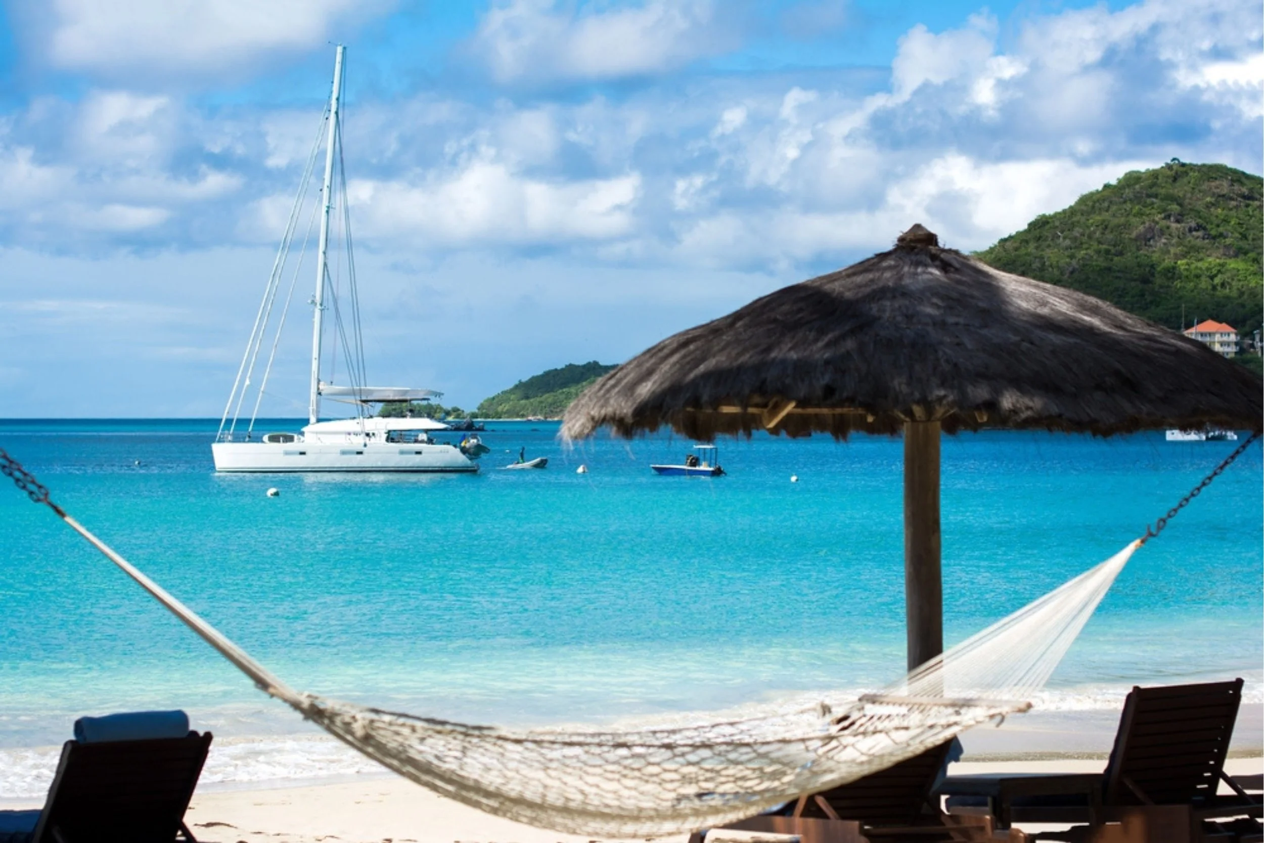 A tropical beach scene with a thatched umbrella, a hammock, lounge chairs, and turquoise waters with boats, a green hillside in the background, and partly cloudy sky.