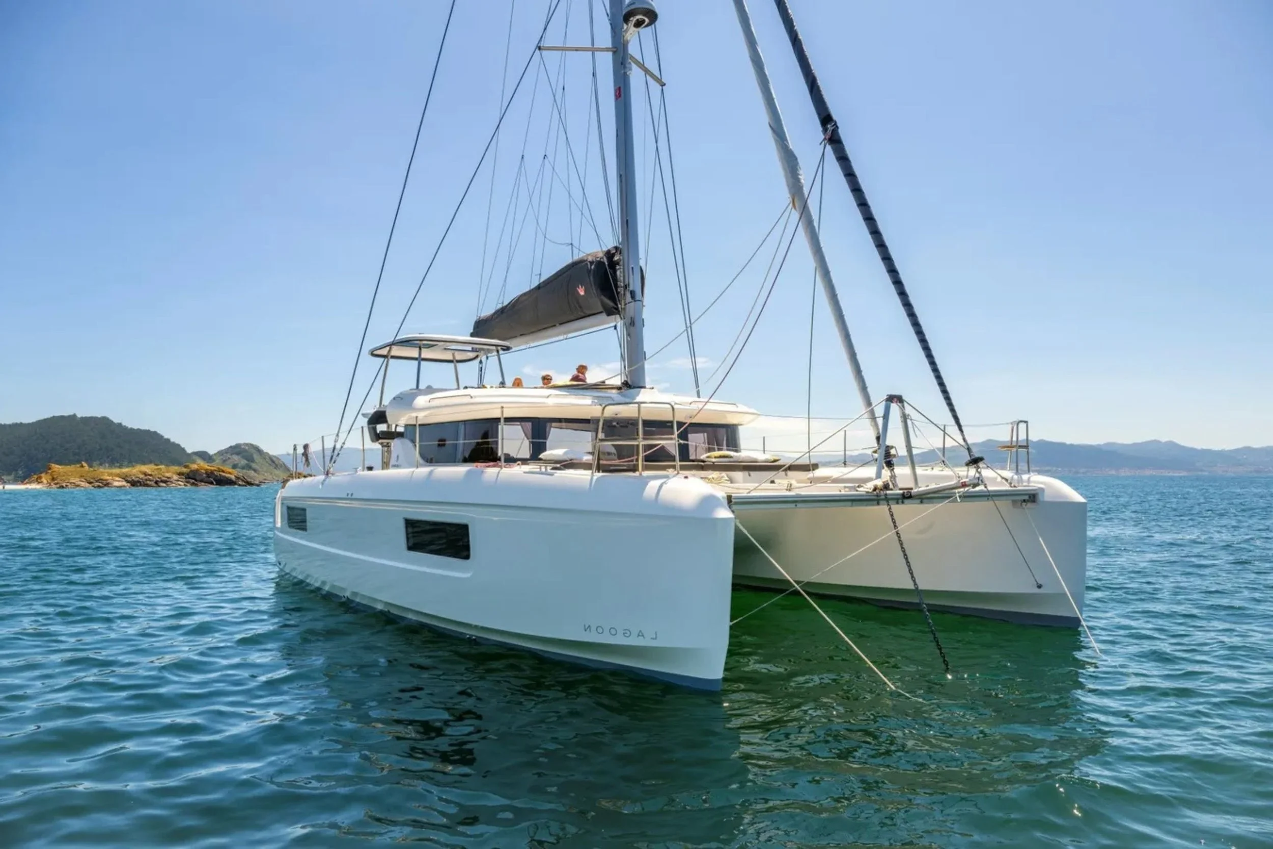 A white sailing yacht anchored on calm blue water with a distant shoreline and mountains in the background under a clear sky.