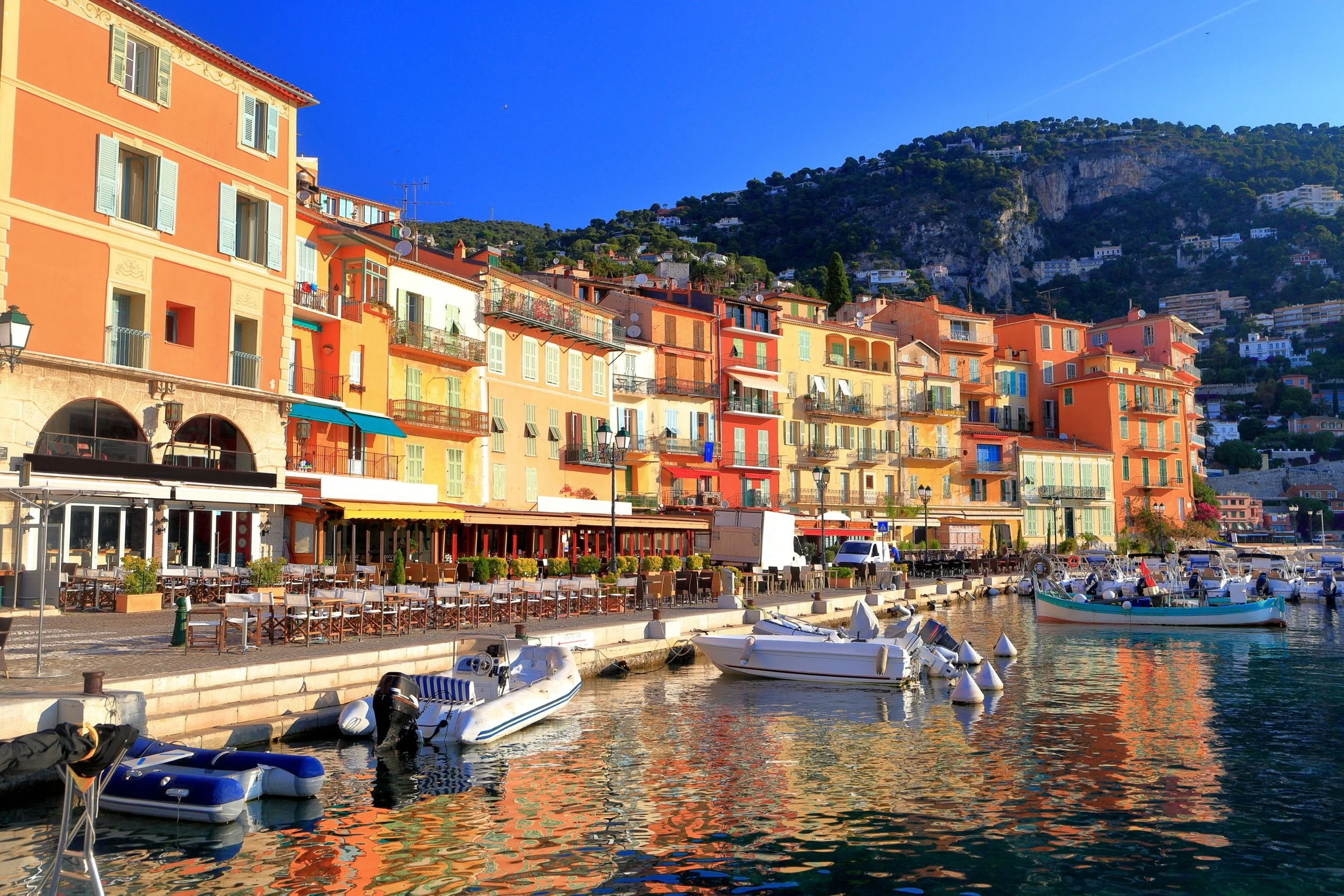 Colorful buildings lining a waterfront with boats docked in the water and a hillside in the background.