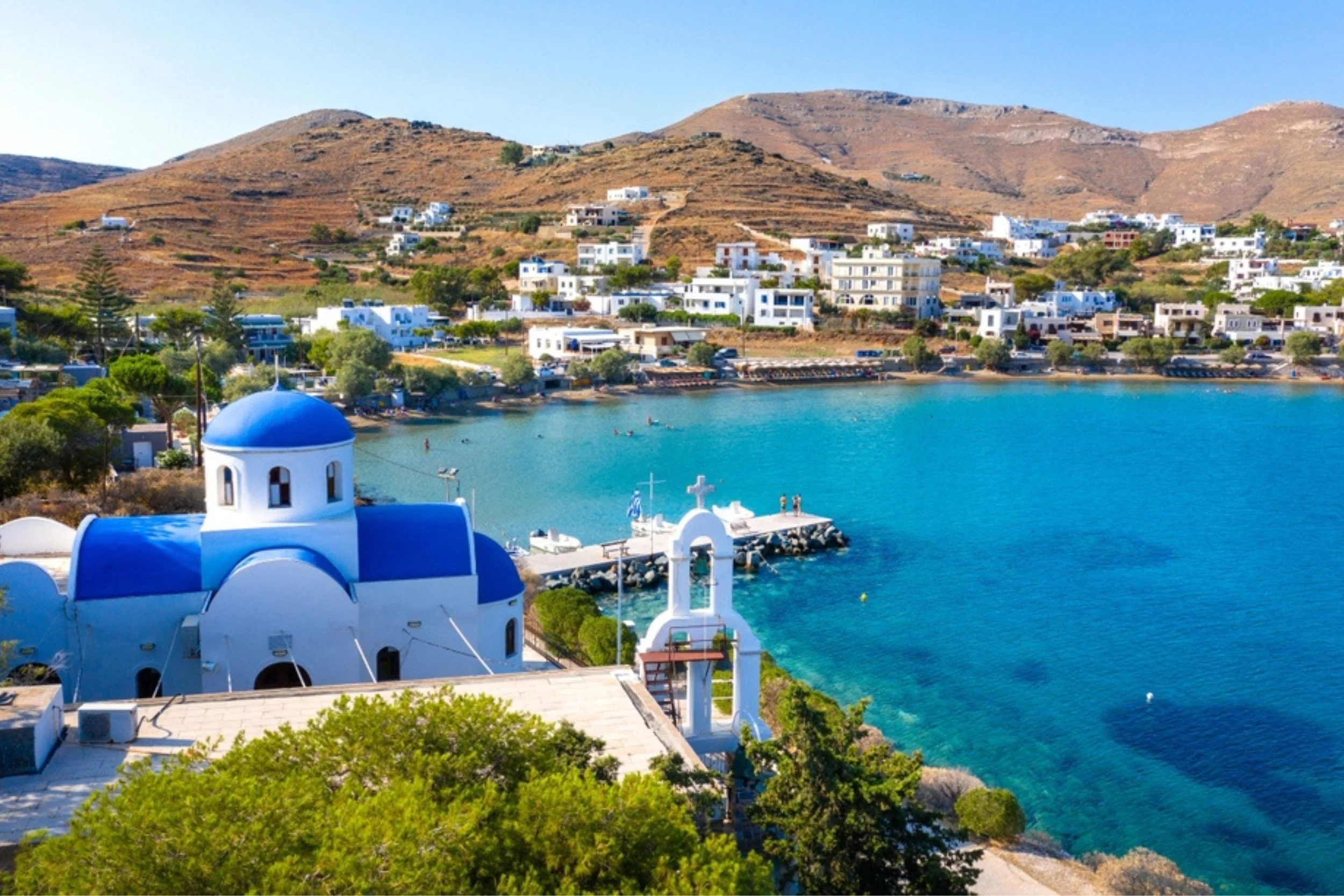 Greek church with white walls and a blue dome on a coast with a small pier and water, hills with white buildings in the background.