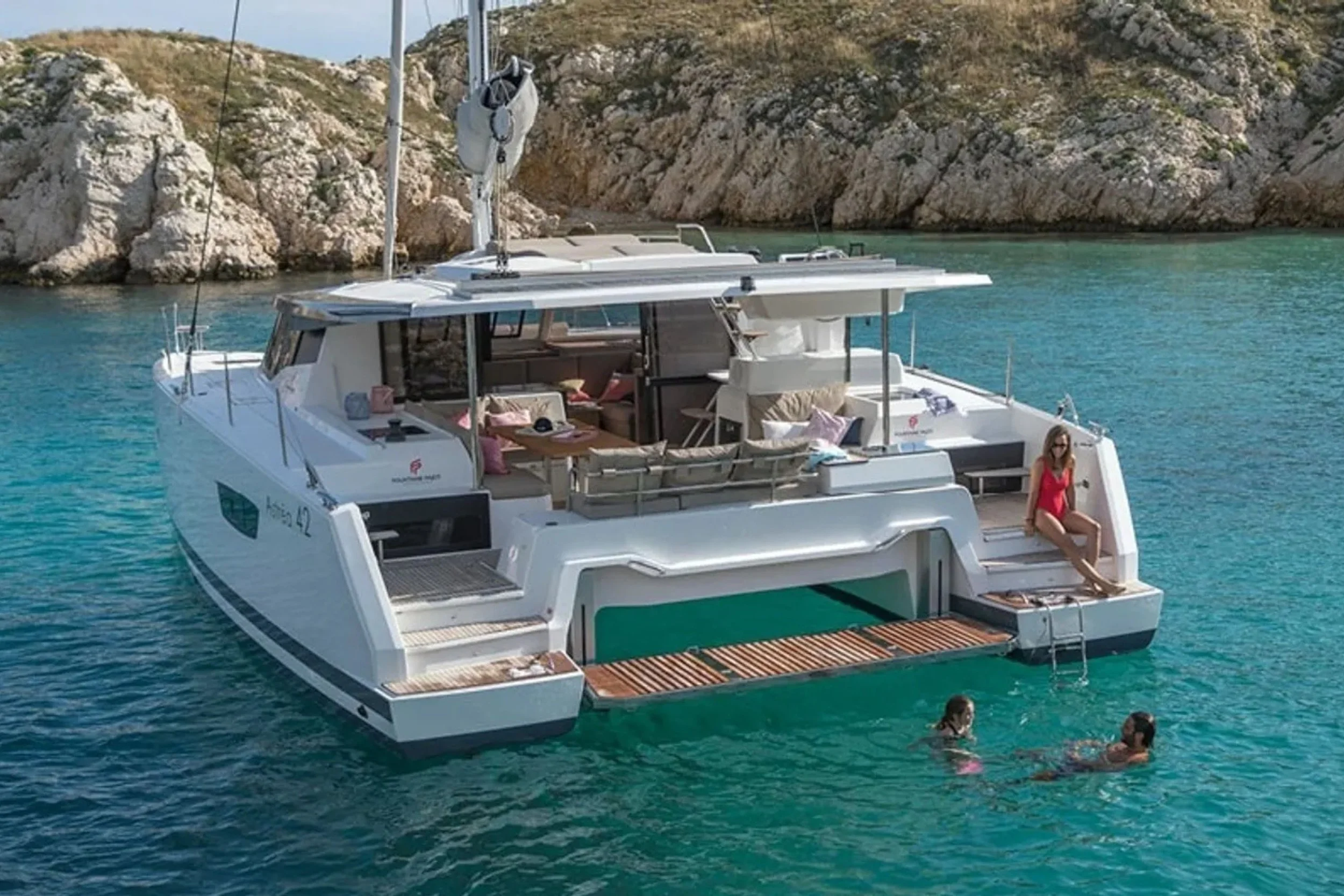 A white luxury yacht anchored in clear blue water near rocky cliffs, with a woman in a red swimsuit sitting on the rear steps and two children swimming nearby.